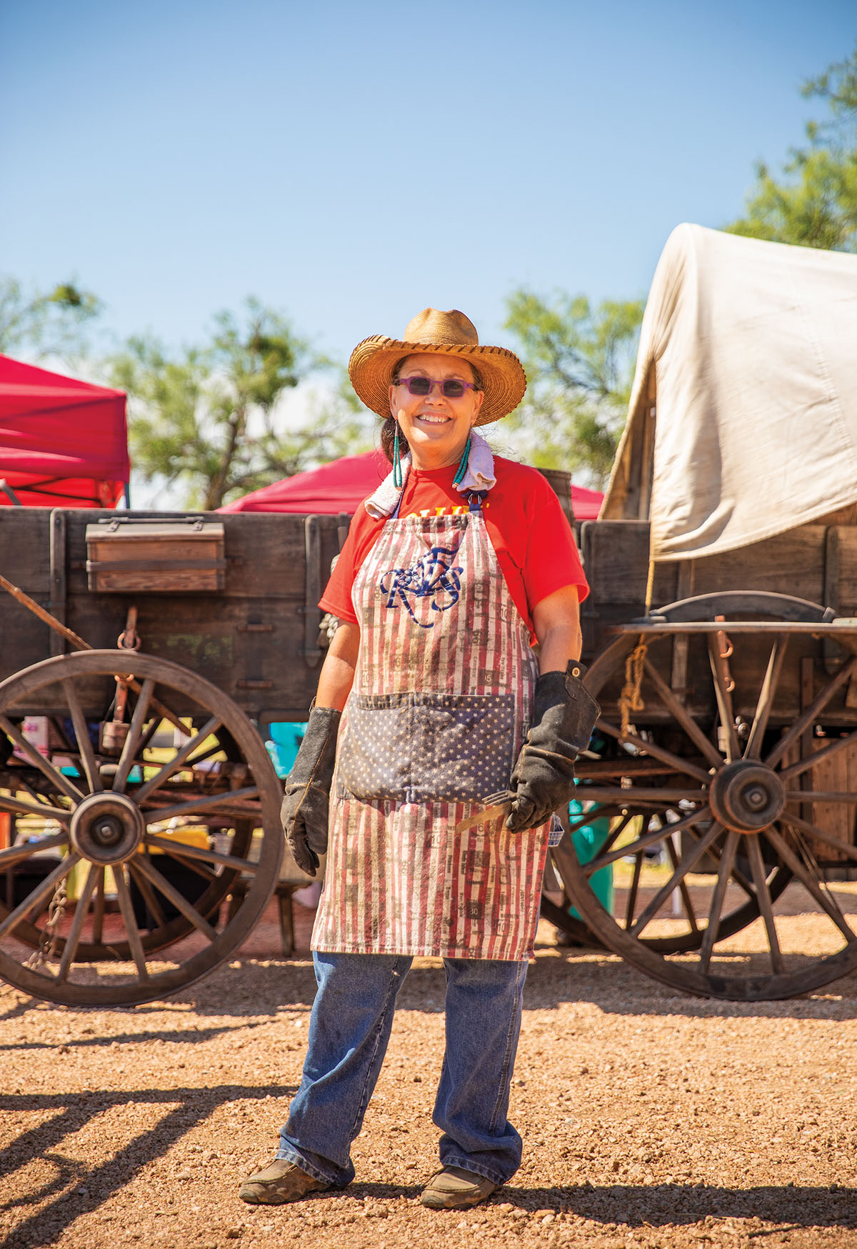 A woman in a sun hat and sunglasses and red shirt and apron stands in front of a traditional wooden wagon