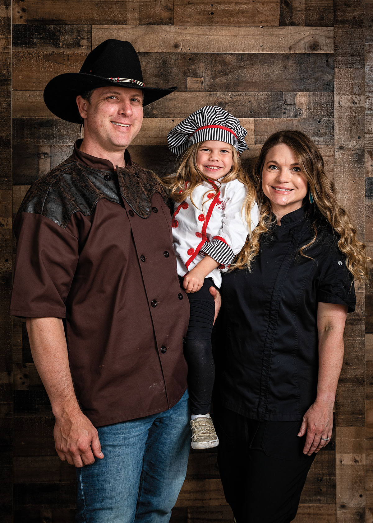 A man in a brown shirt and cowboy hat and woman in black chef's uniform hold a child in a white chef's uniform and checkered hat