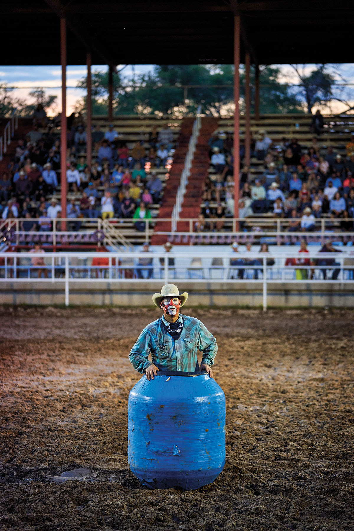 A man in a bright blue shirt, face makeup, and a white hat sits in a large blue barrel in front of a crowd of onlookers