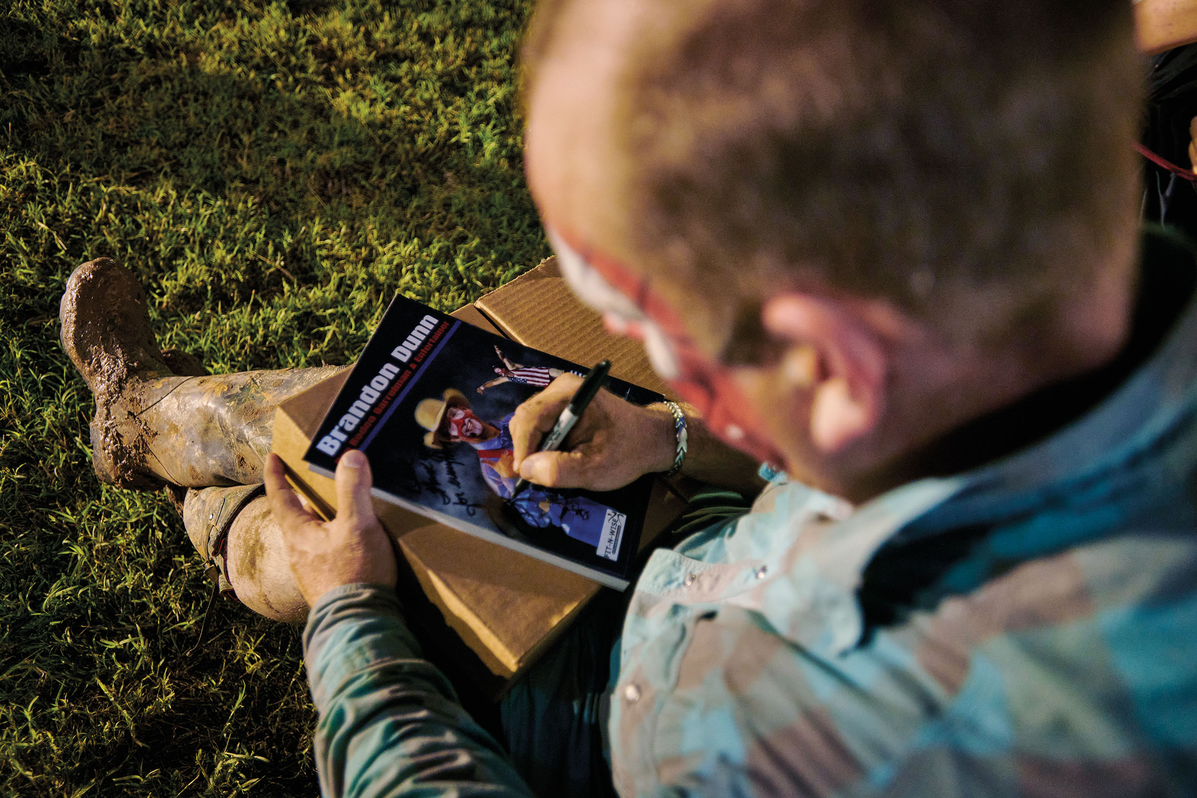 A man in rodeo clown facepaint sits on the grass signing a small poster