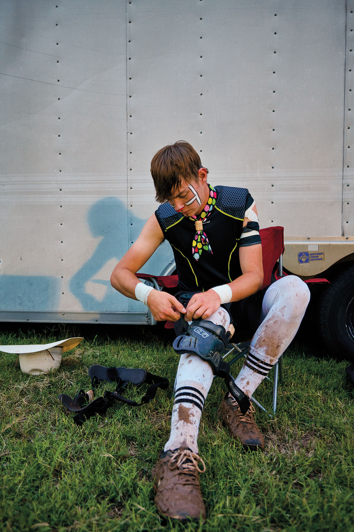 A young man puts on protective equipment in front of a white trailer on green grass