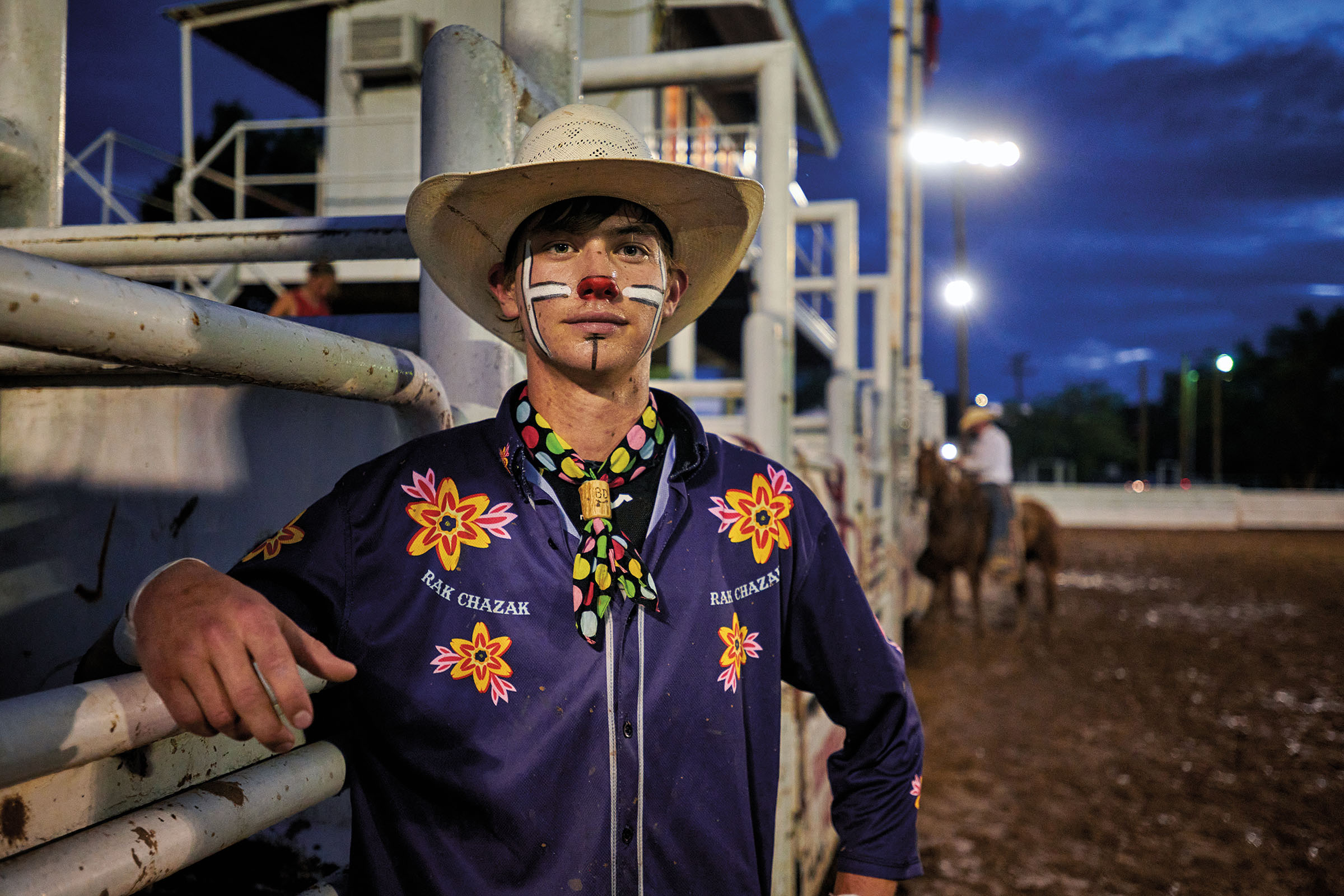 A man with a tan hat, face paint including a red nose and white stripes, and a bright purple shirt looks at the camera under flourescent lights