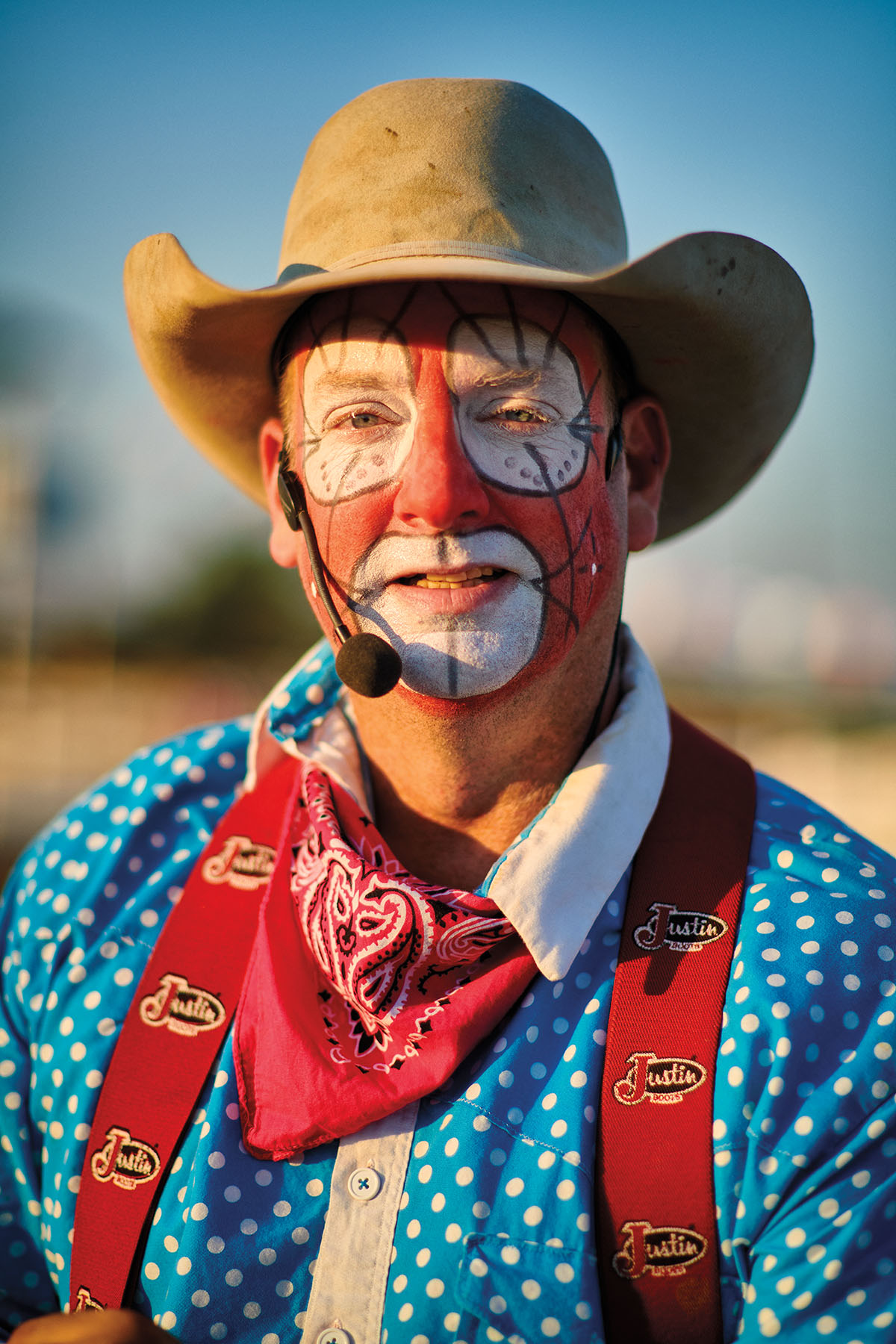 A man in a tan cowboy hat, face makeup, a wireless microphone and a bright blue shirt with red suspenders looks at the camera