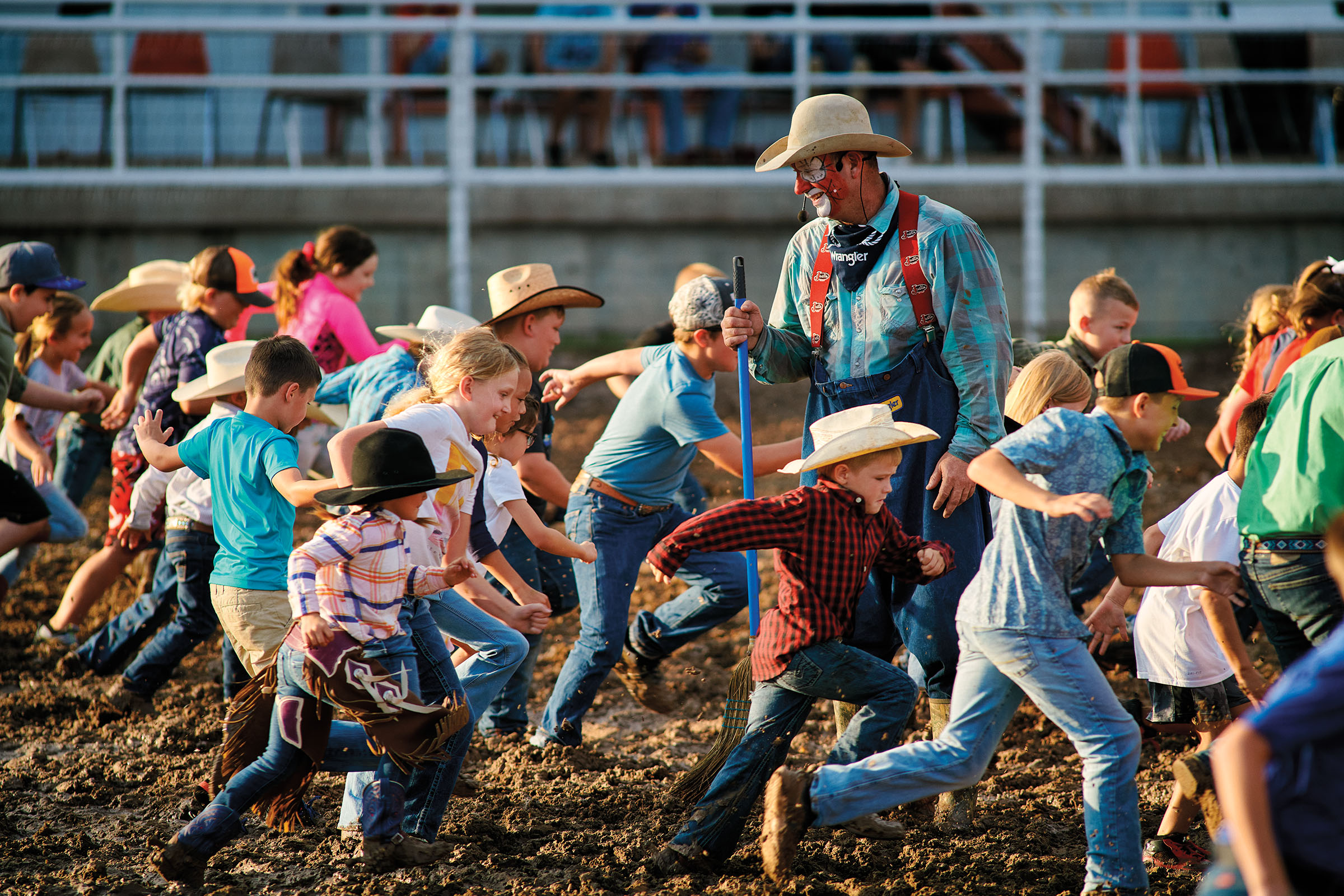 A group of children dressed in bright colors and cowboy hats run past an adult rodeo clown standing up