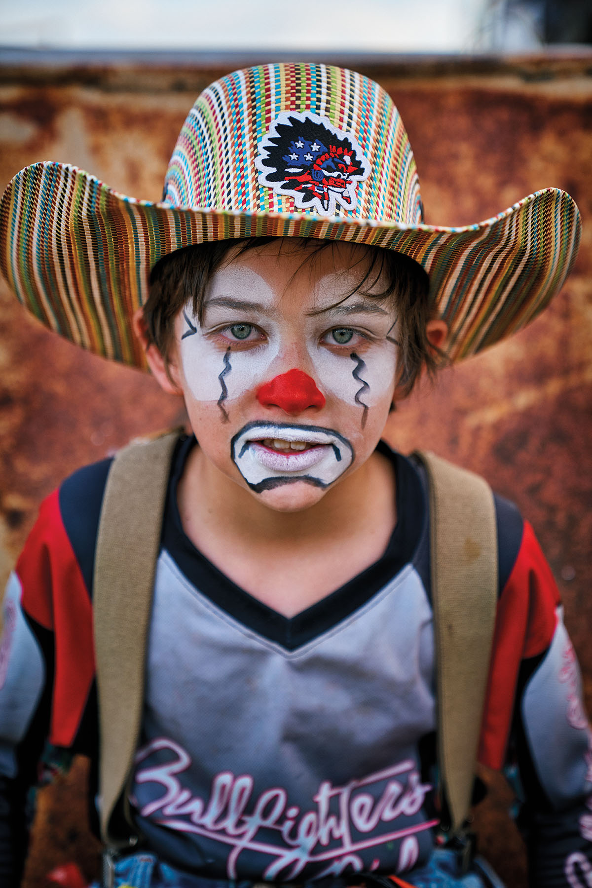 A young man in a striped cowboy hat, white paint around his eyes and mouth, a red nose, and a bright red and gray shirt looks into the camera