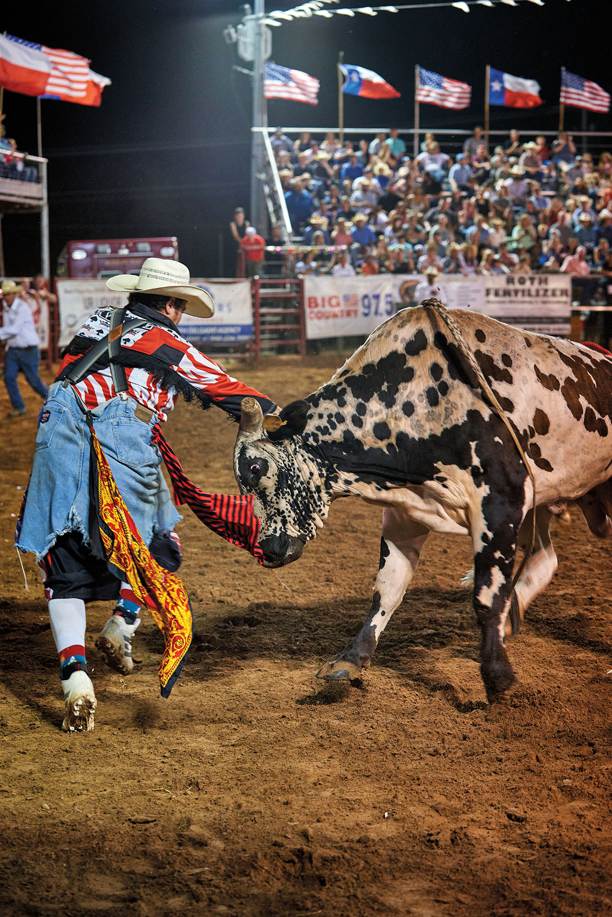 A man dressed in bright red, blue and yellow flowing clothes and a white cowboy hat holds his arms out to a bull