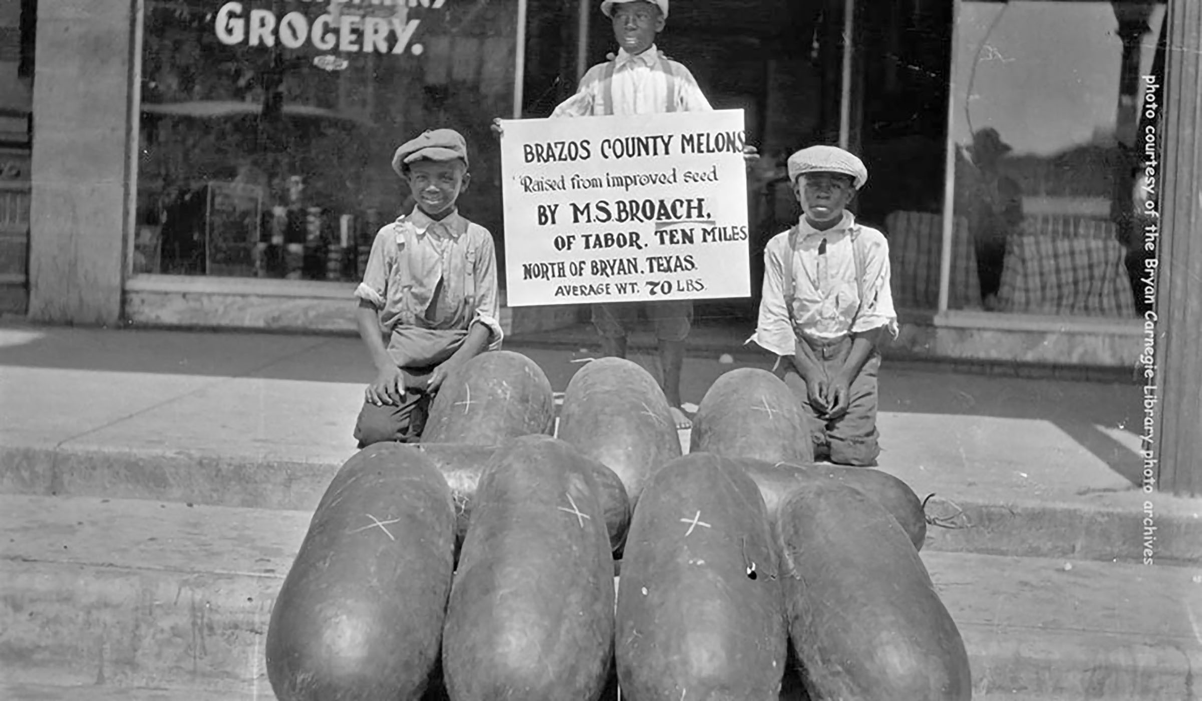 Three boys sit in front of a stack of very large watermelons