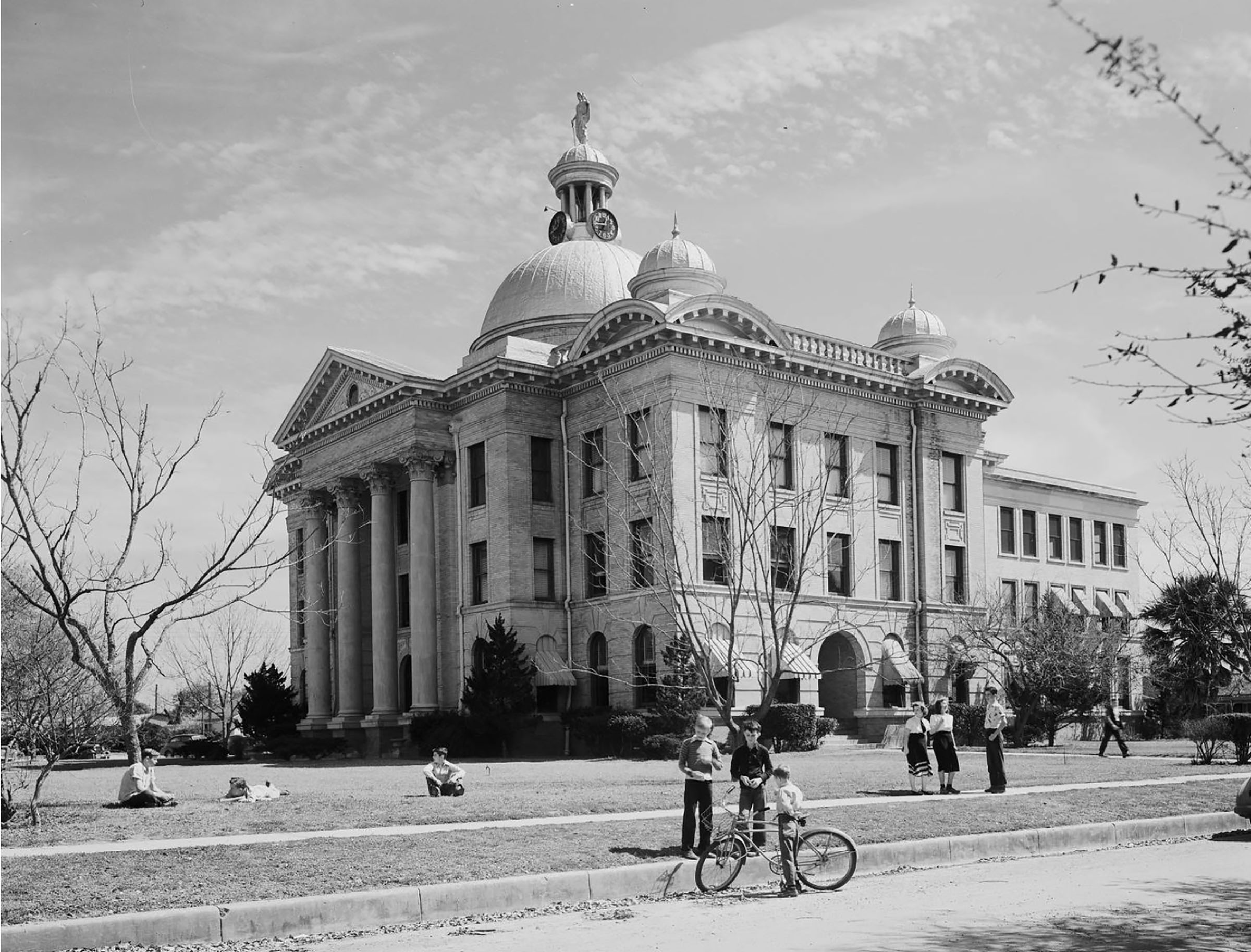 Children stand with bicycles and mill about on the grass in front of a tall stone courthouse