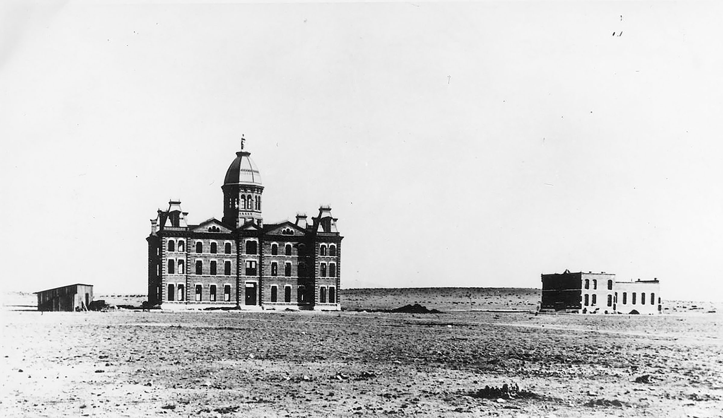 A three-story building with an ornate tower sits in the middle of the desert, with mountains visible in the background