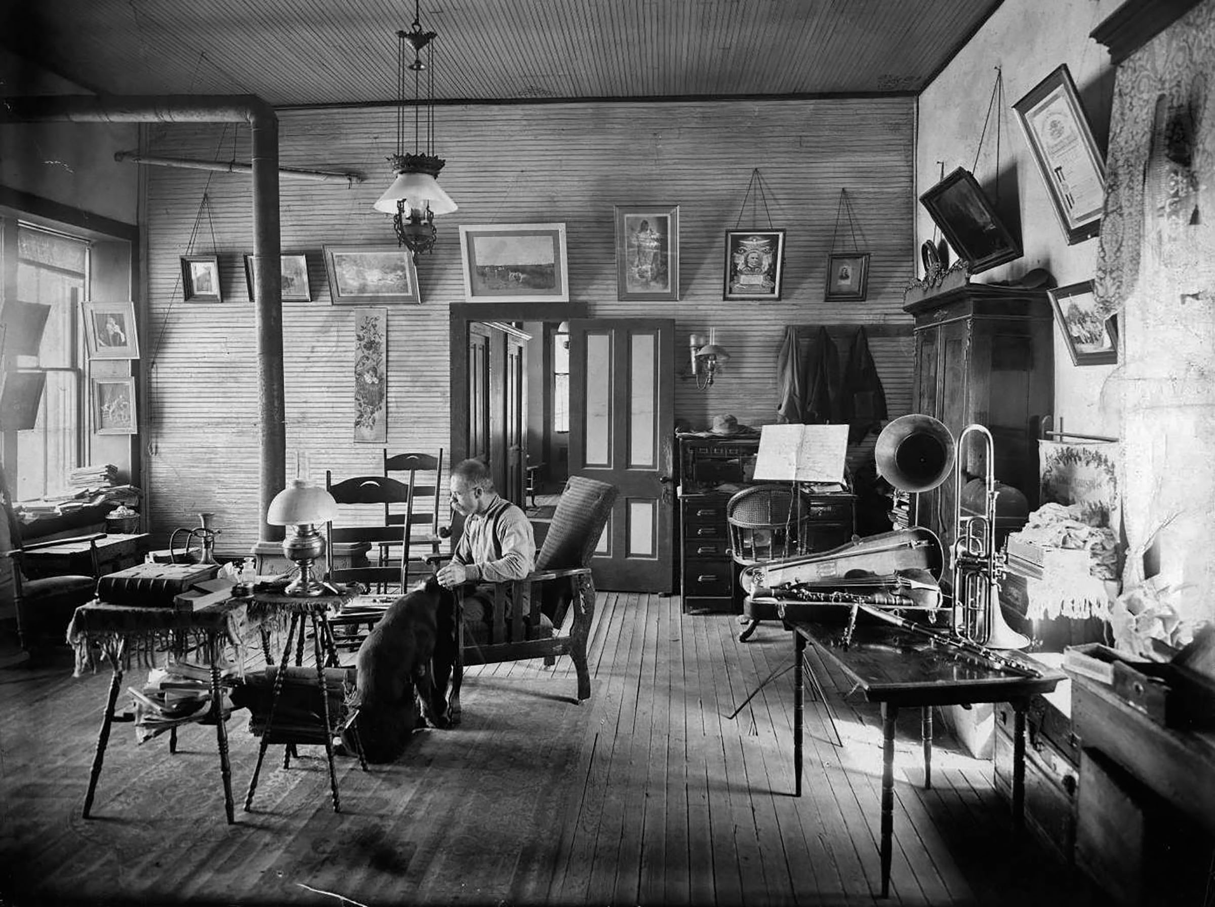 A man sits in a well-furnished wooden room with a photograph, chairs and paintings