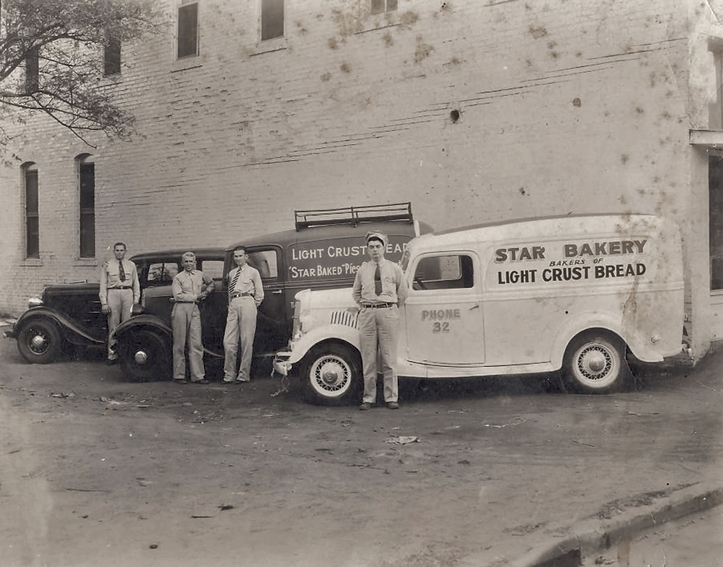 Men in dress shirts and ties stand in front of bakery vehicles and a brick wall