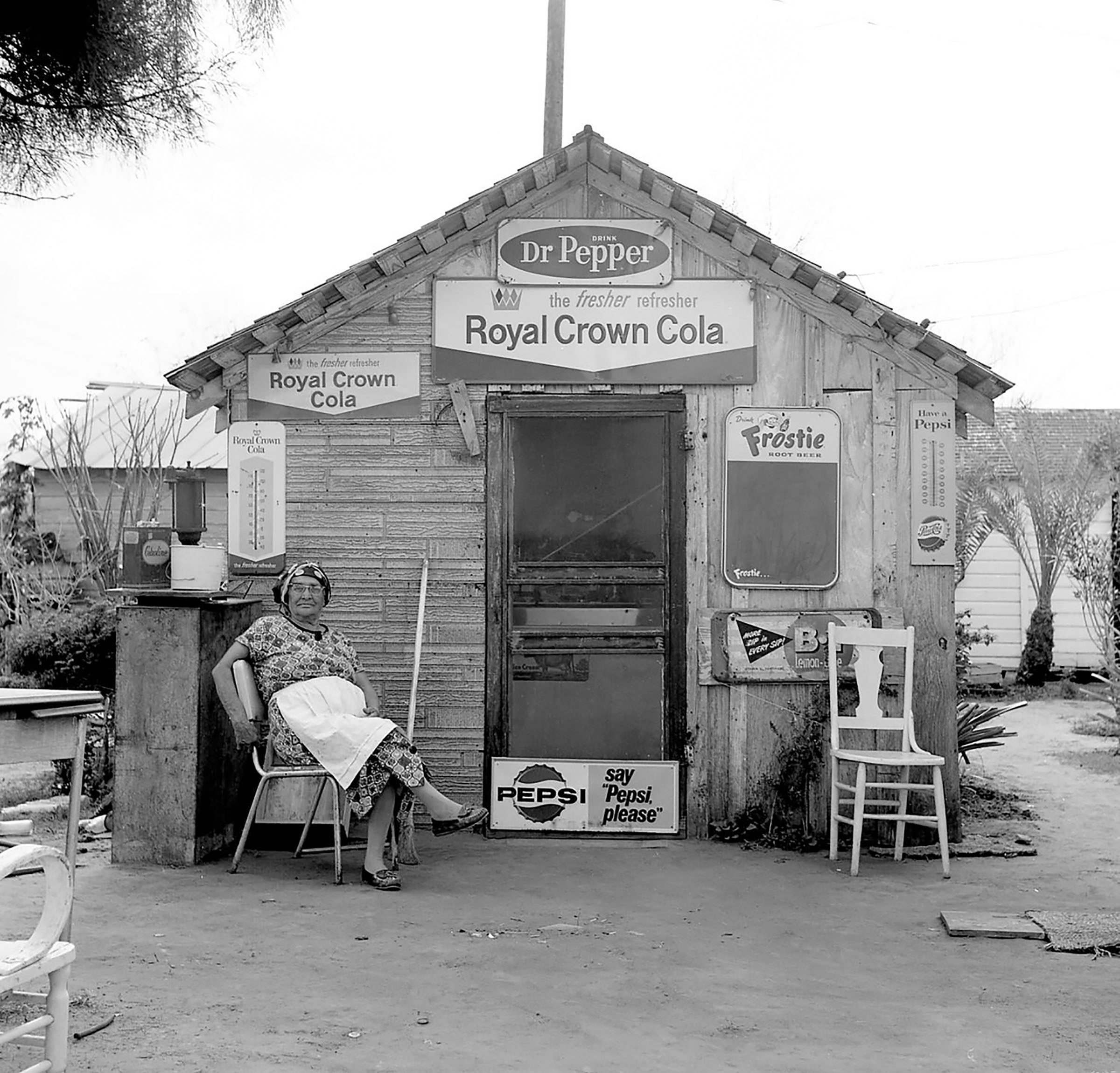 A woman in a white apron sits in front of a wooden storefront with signs advertising Dr Pepper, Royal Cola and Pepsi
