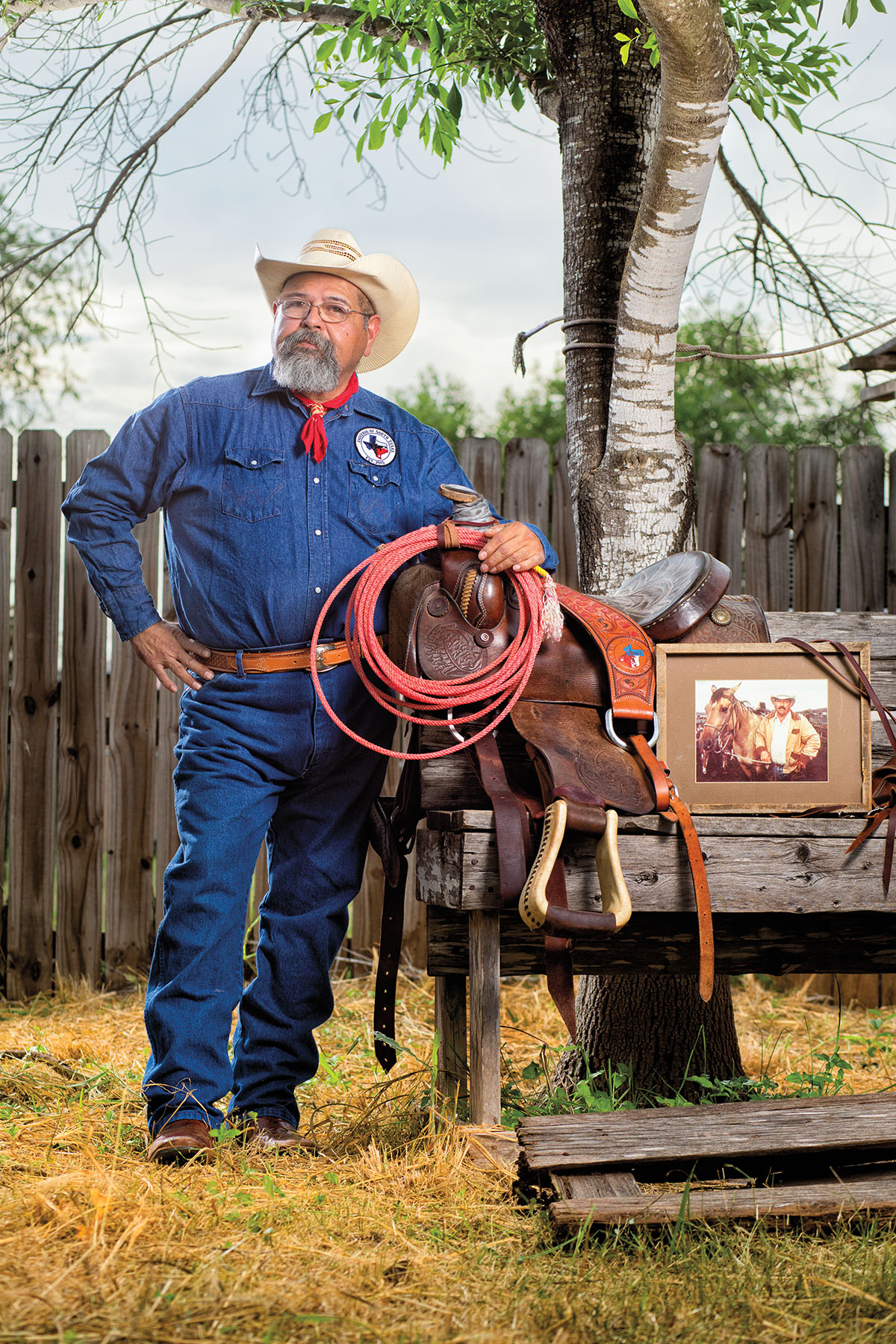 A man in white cowboy hat, blue shirt and blue jeans holds a rope next to a vintage photograph outdoors