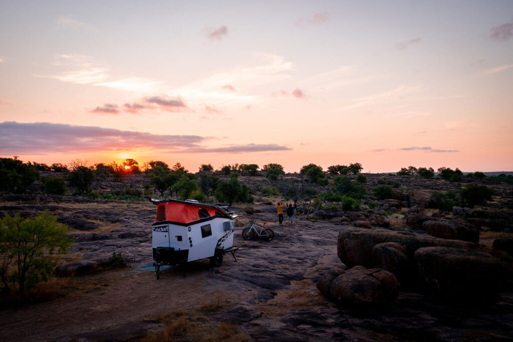 A TAXA Cricket camper parked on a flat, rocky landscape with a sunset in the distance. 