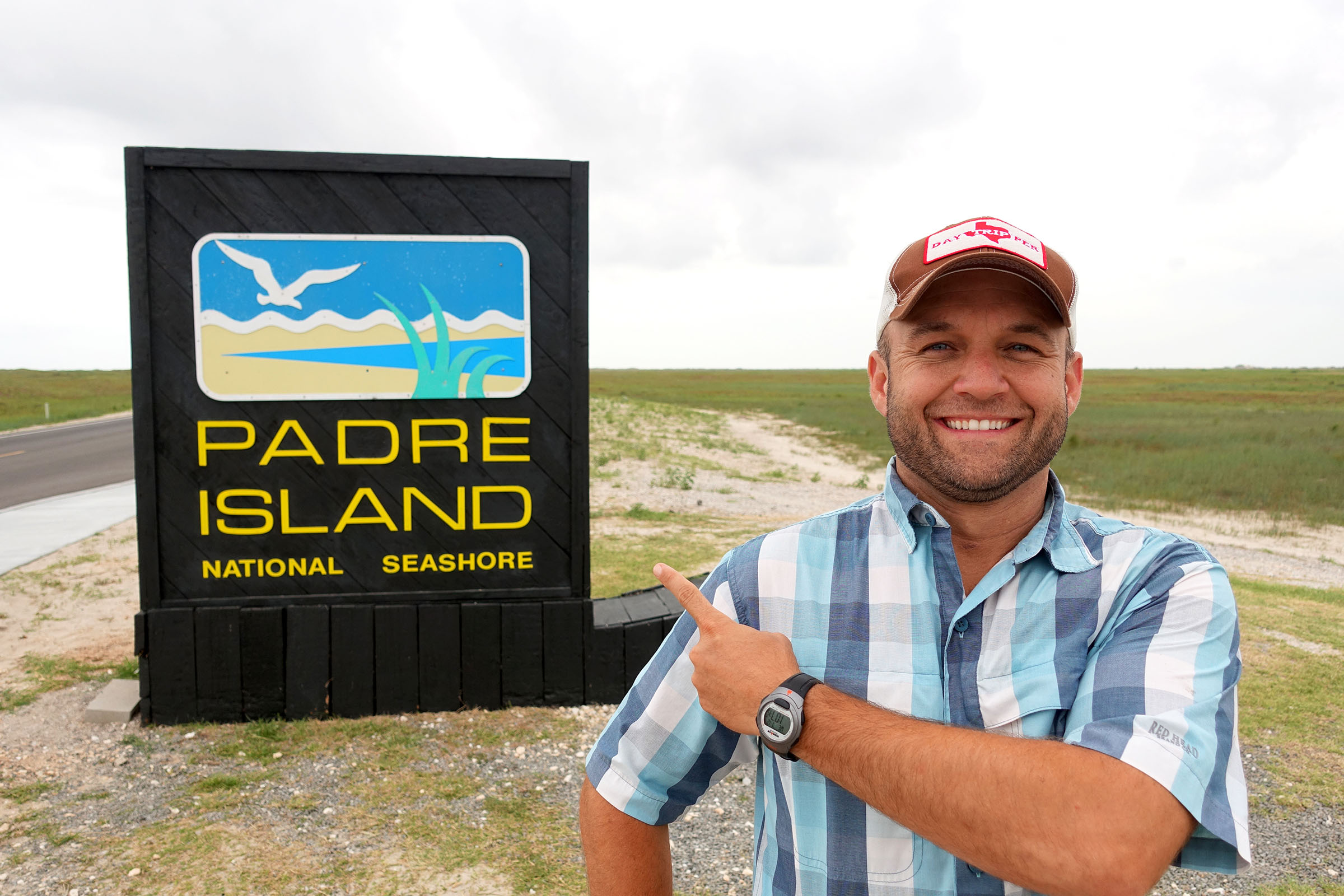 A man in a plaid shirt and baseball cap smiles and points at a sign reading "Padre Island National Seashore"