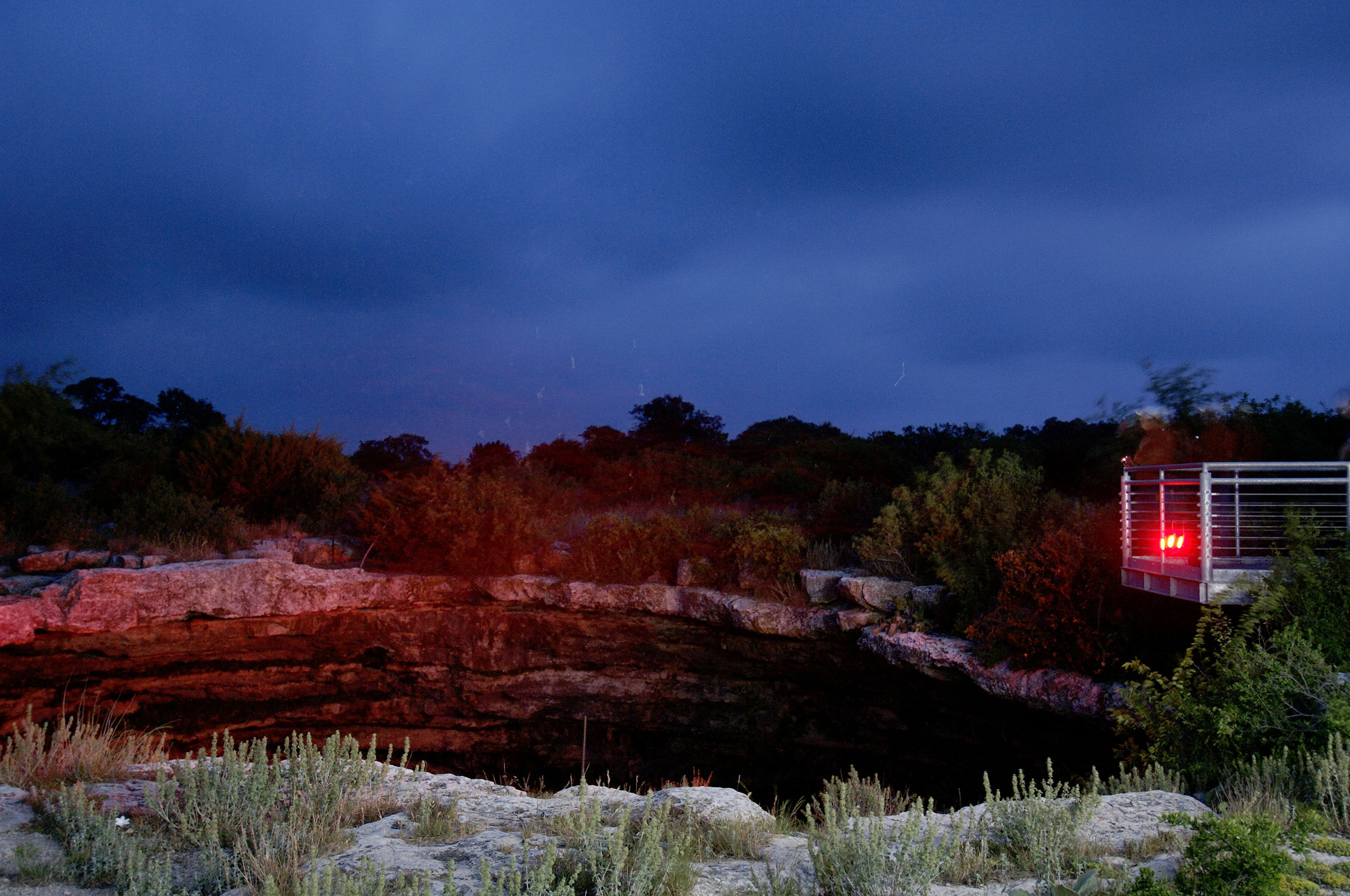 Red lights illuminate the side of a large hole in the limestone surrounded by short plants, under a dark blue dusk sky