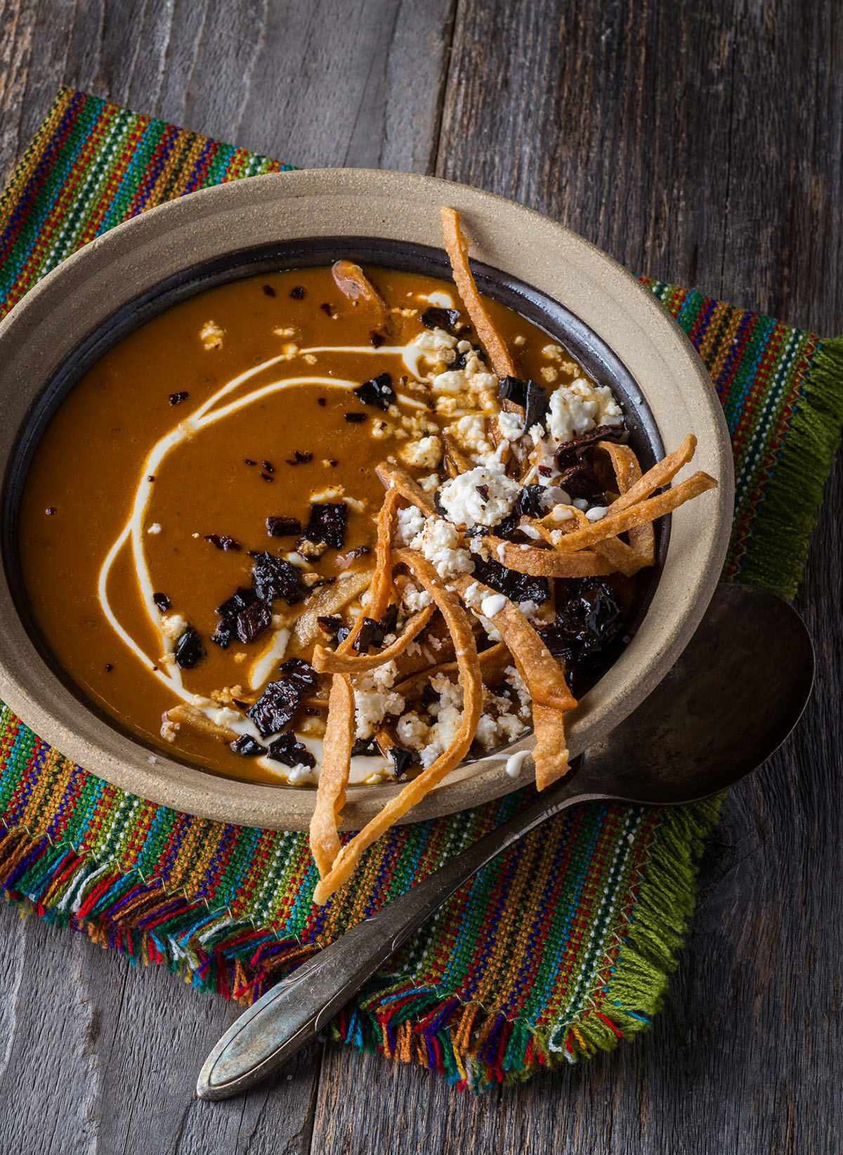 A side view of brown soup topped with black beans, cheese and tortilla stips in a stoneware bowl on a wooden background