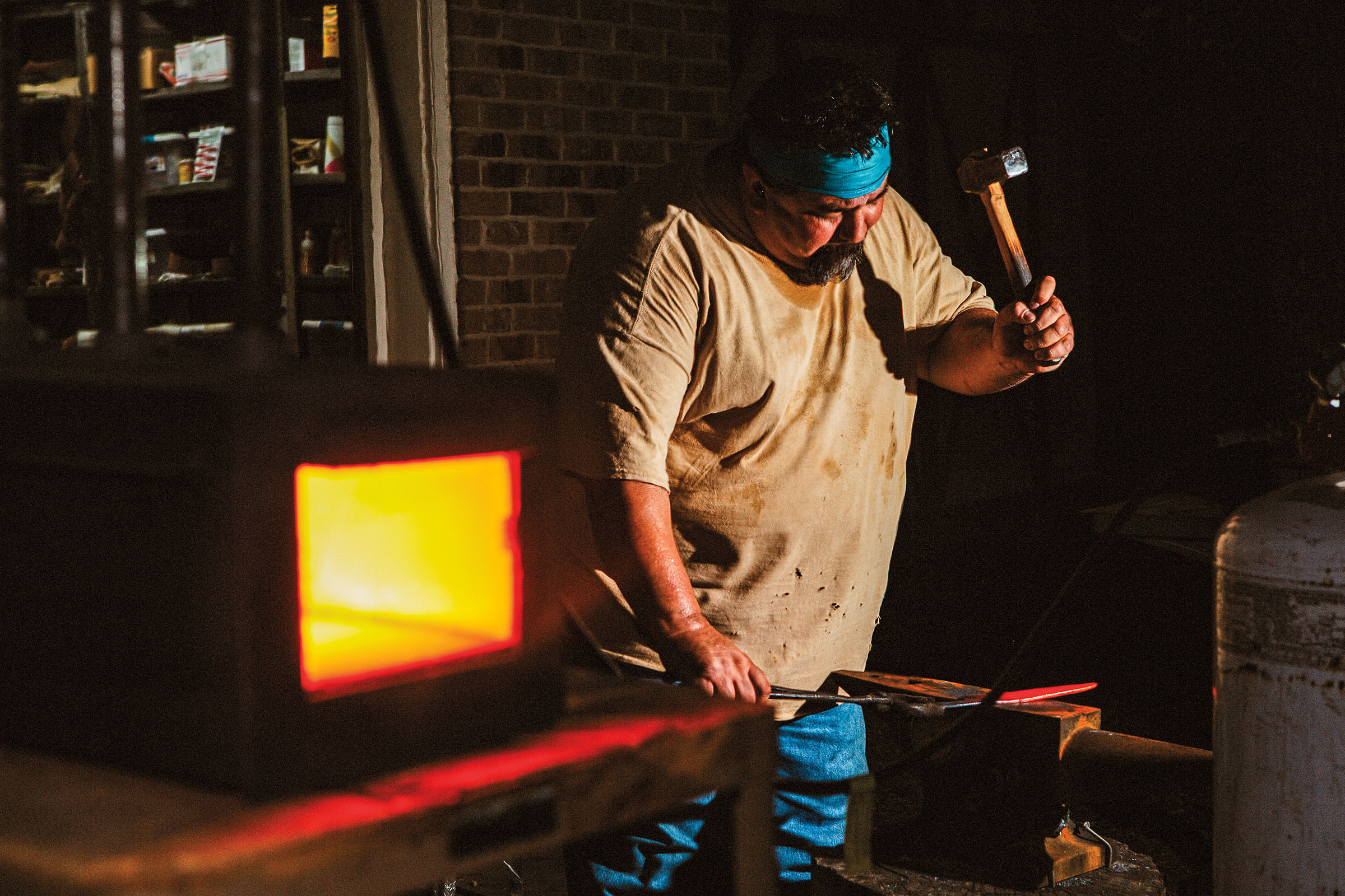 A man in a tan shirt and turquoise headband holds a hammer above his head in front of a bright firebox and glowing steel