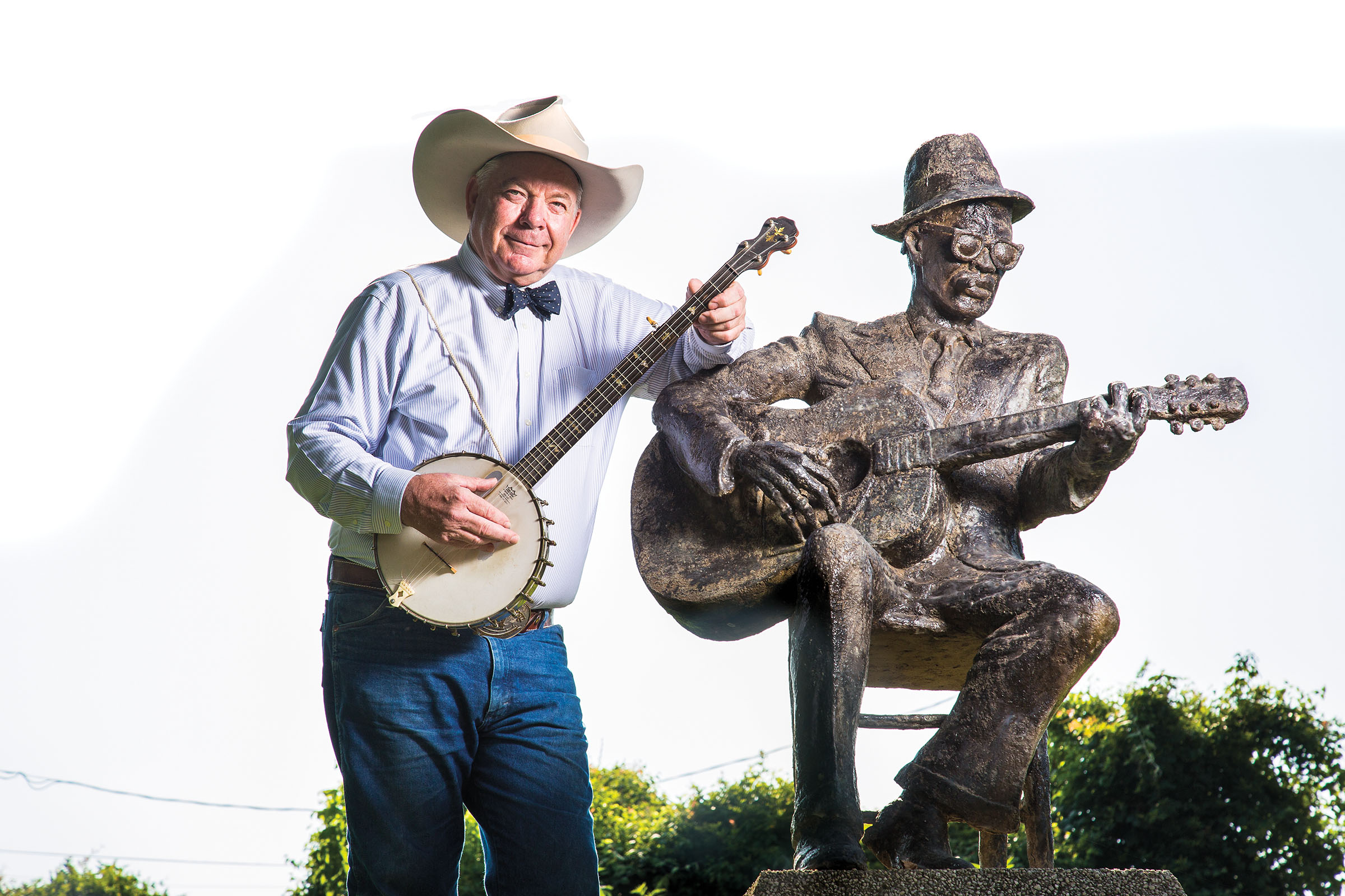 A man in a cowboy hat holding a banjo leans on a statue of a man in a small hat holding a guitar