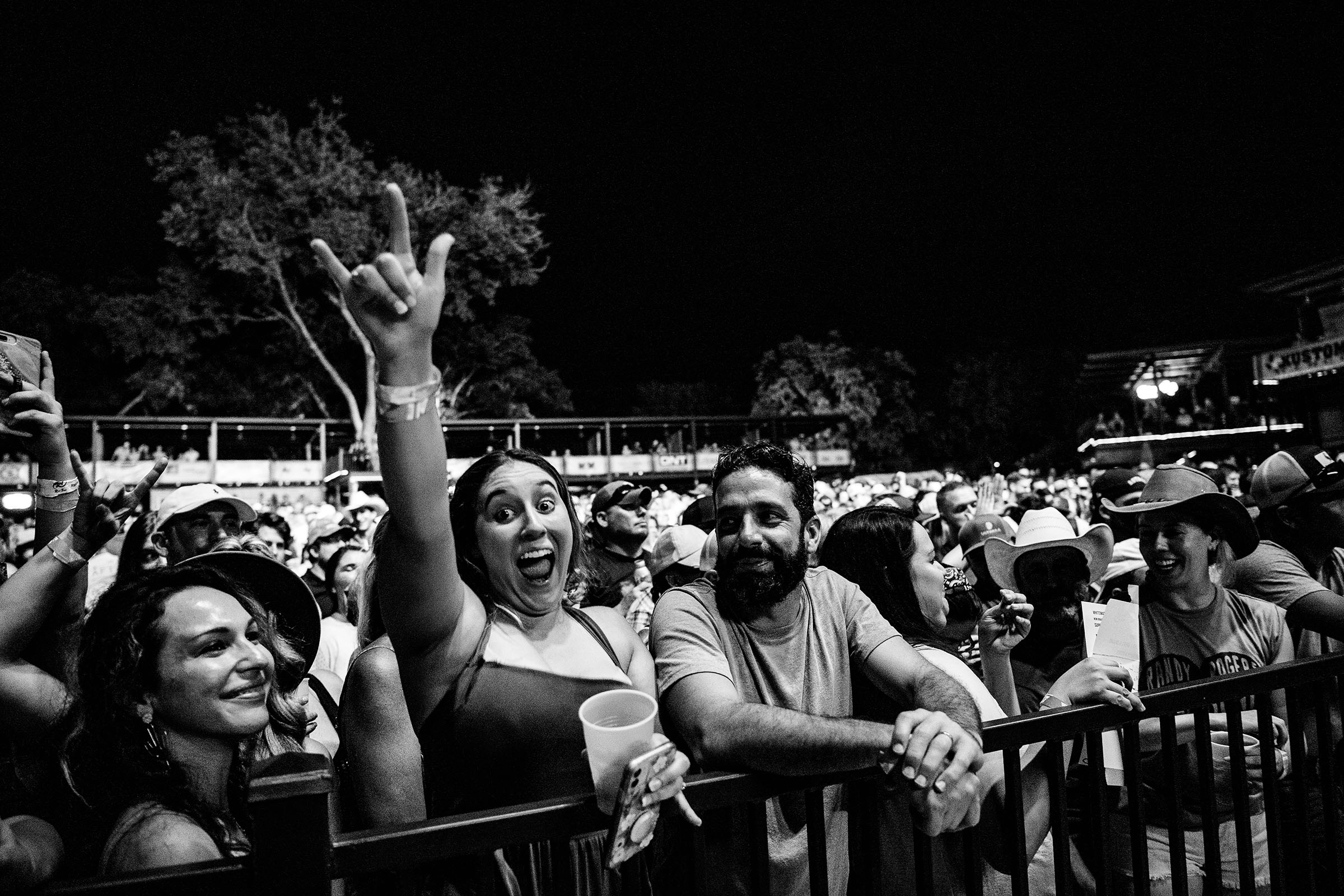 A woman puts her hand up in the air with a large smile in a group of people attending a concert