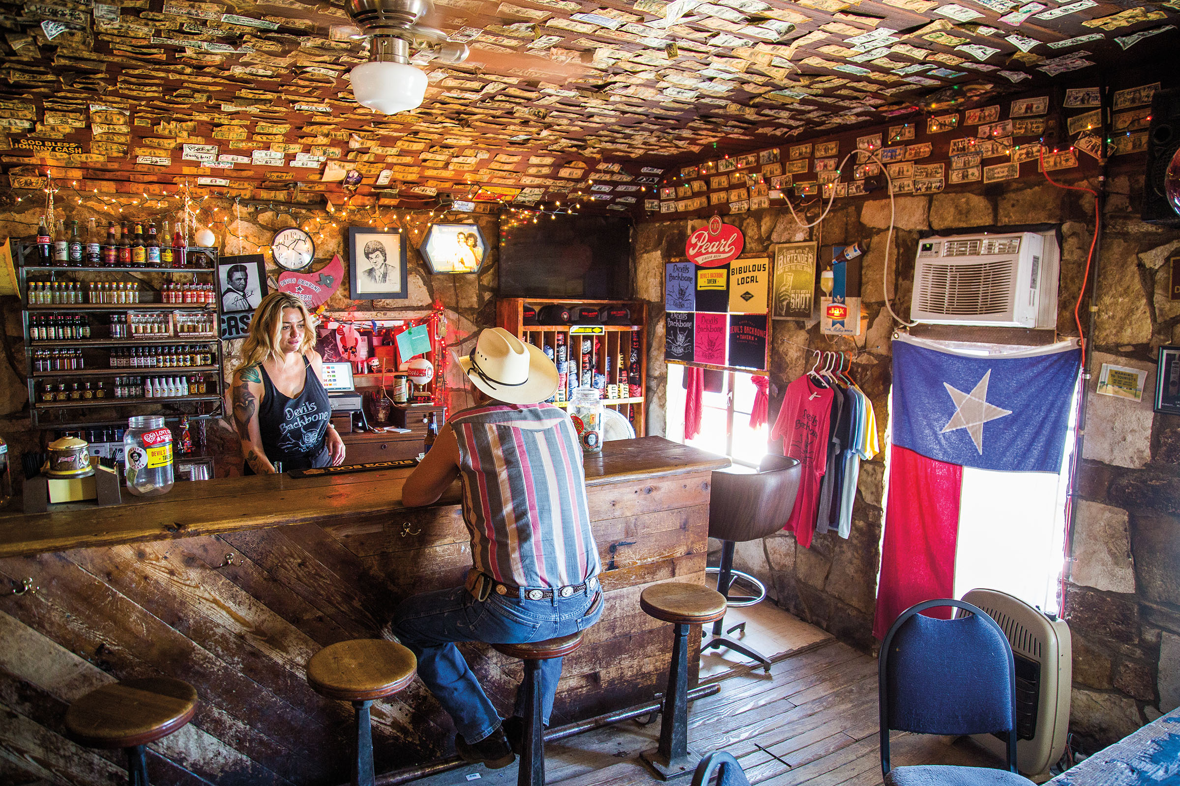 A man in a cowboy hat sits at a heavily-decorated bar talking to a female bartender. A Texas flag covers the window, and dollar bills line the ceiling.