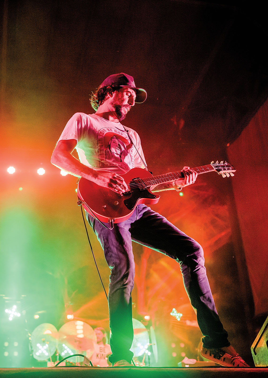 A man in a baseball cap strums an electric guitar and taps his foot on a stage in front of orange and green gel lights