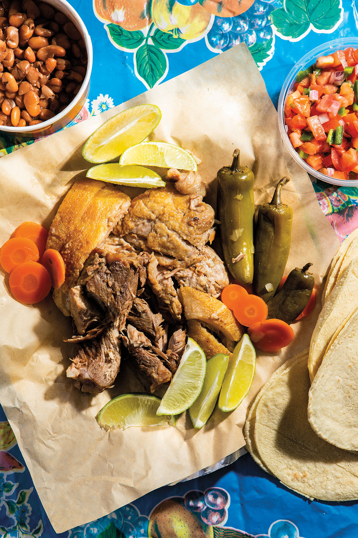 An overhead view of a platter of carnitas with lime wedges, carrots, jalapenos and corn tortillas