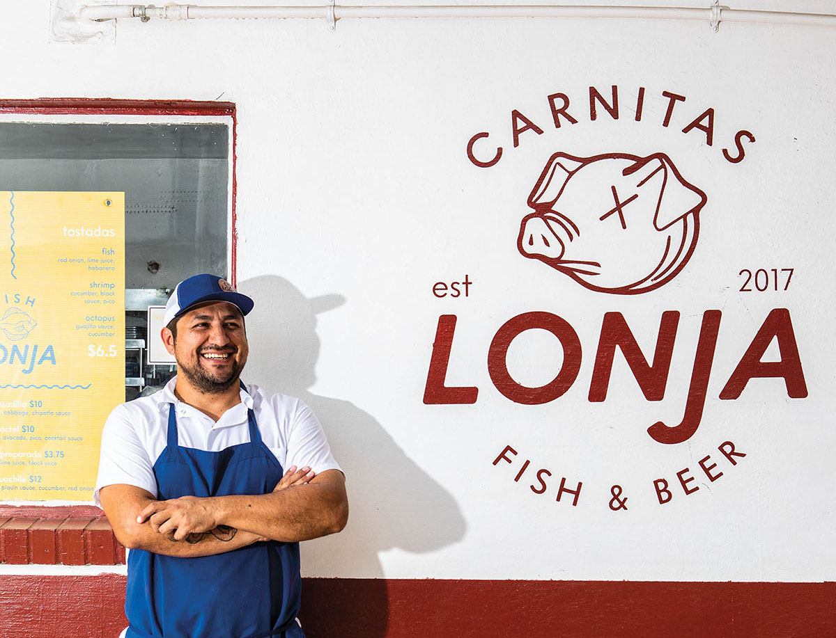 A man in a blue apron and ball cap stands with his arms crossed in front of a red and white truck reading "Carnitas Lonja Fish & Beer"