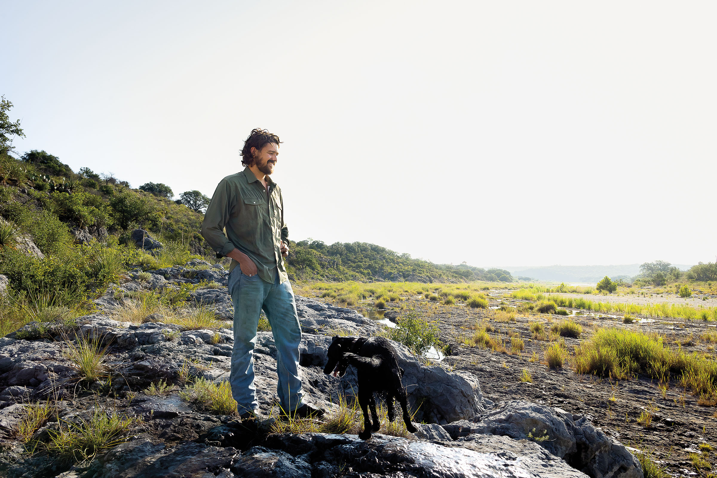 A man smiles and tucks his hands into his pockets while walking through a rocky landscape with a black dog