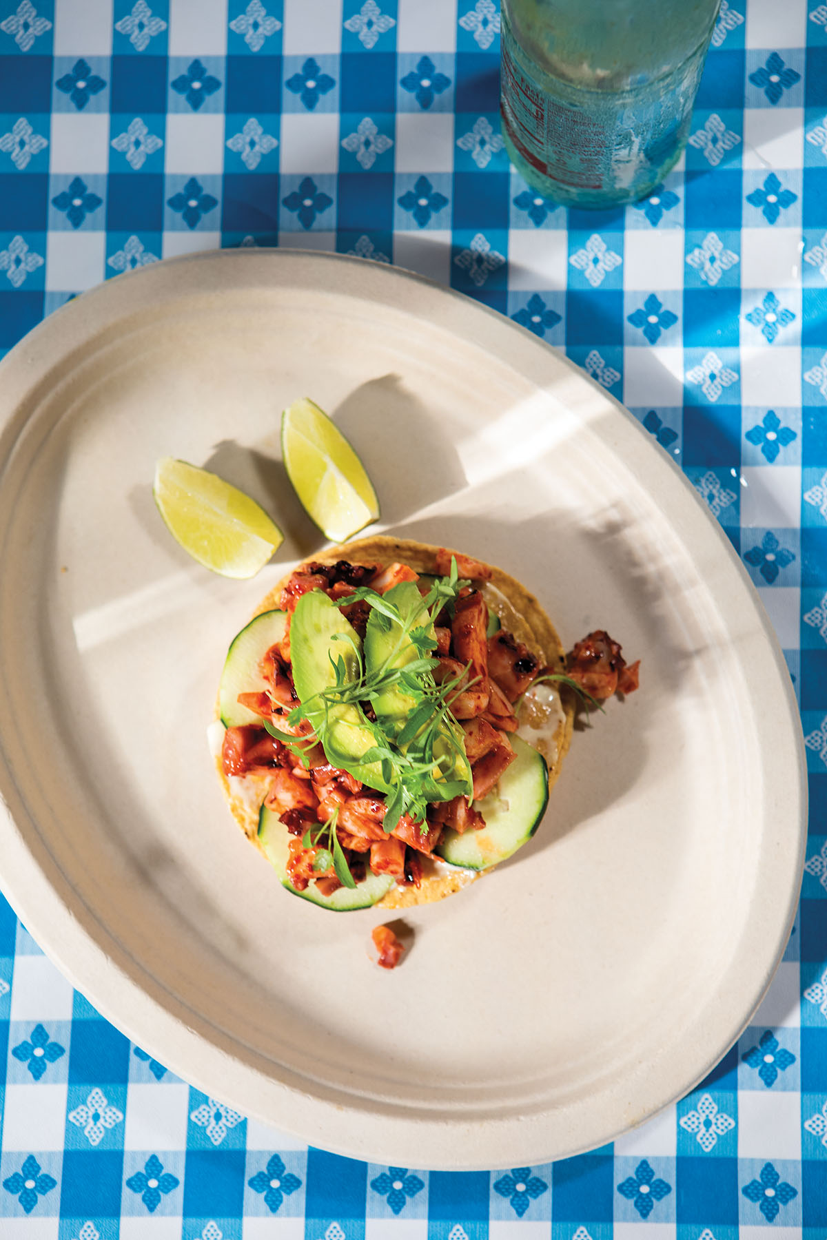 An overhead view of a blue and white checkered tablecloth and a large paper plate holding a taco with bright red topping and avocado