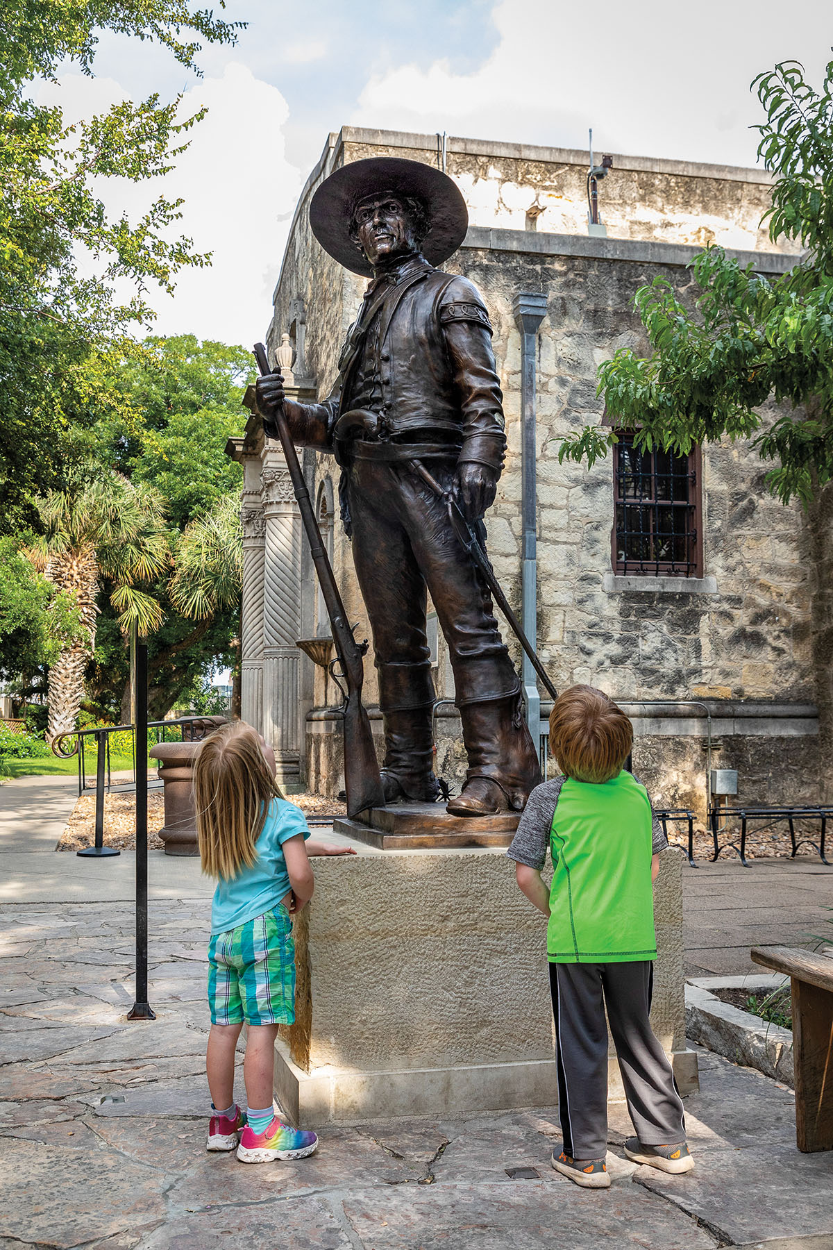 Two childen look up at a dark bronze statue of a man in a large hat holding a sword