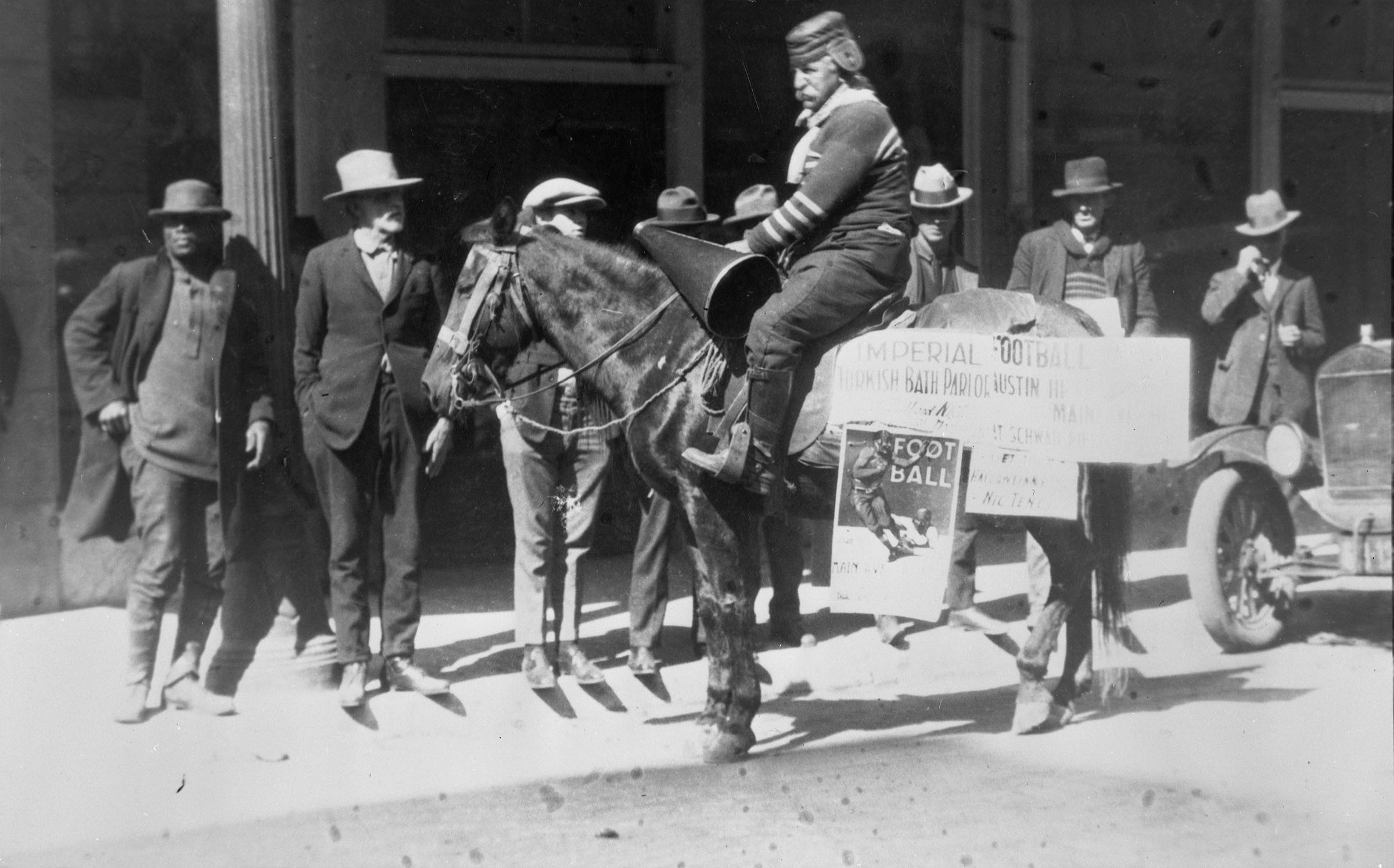 A man in old-timey football gear sits on horseback in front of eight people in a black-and-white photograph