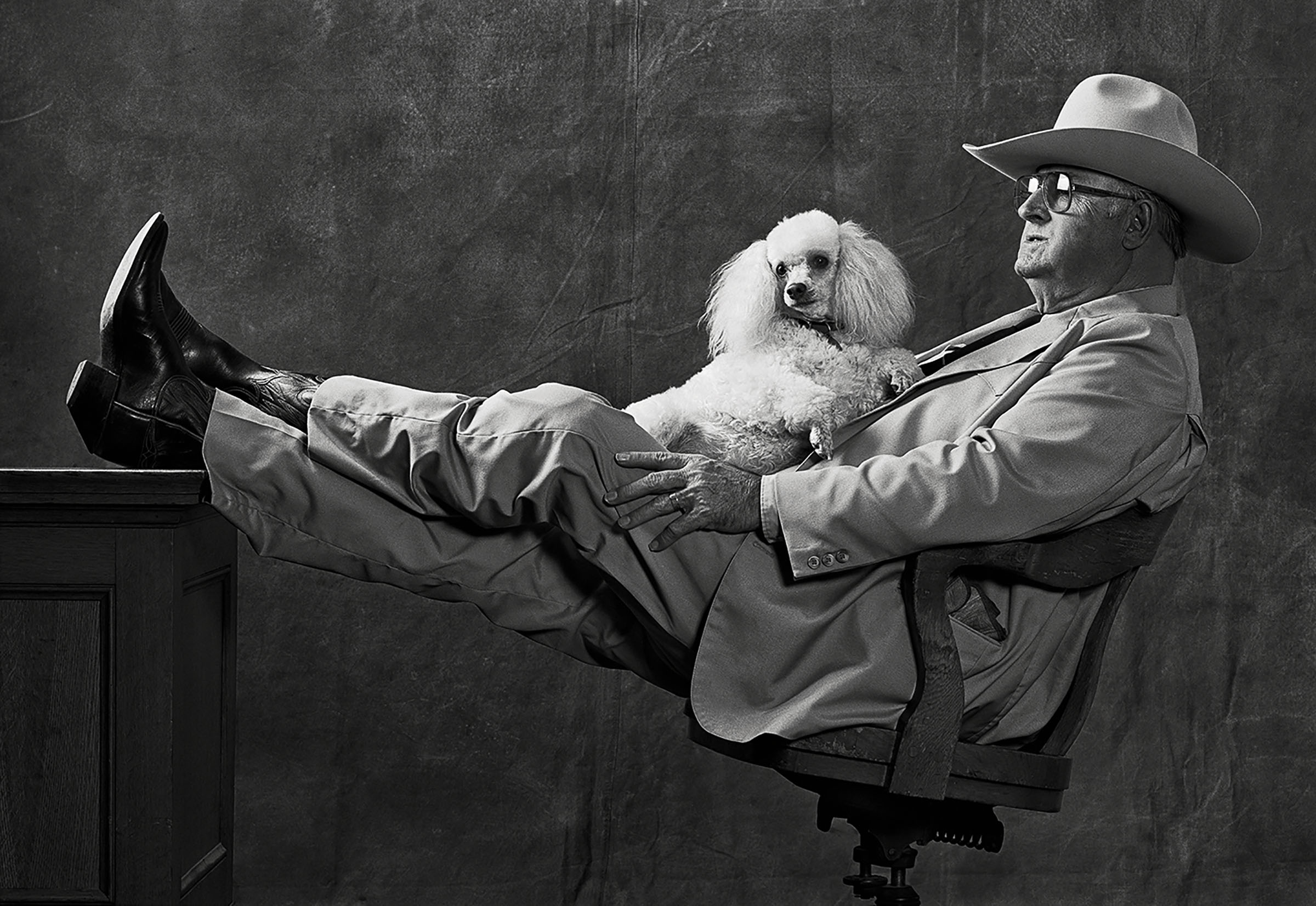 A man in a large cowboy hat kicks back with his feet up on a desk and a small white dog in his lap