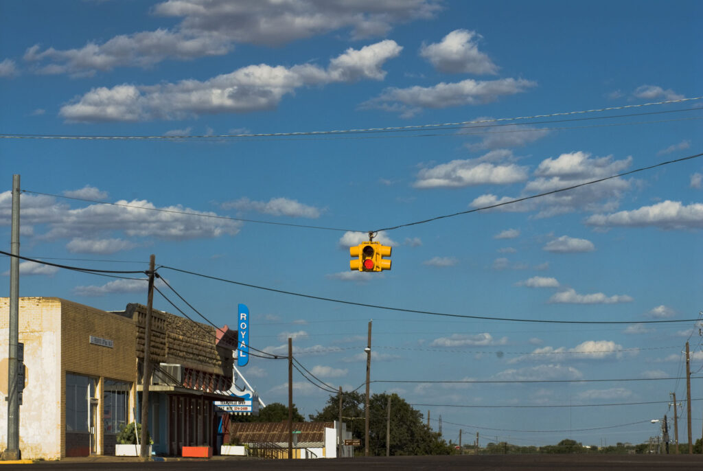 A yellow stoplight hangs from a black wire over a town street, with a building on the left displaying a blue sign reading "ROYAL"