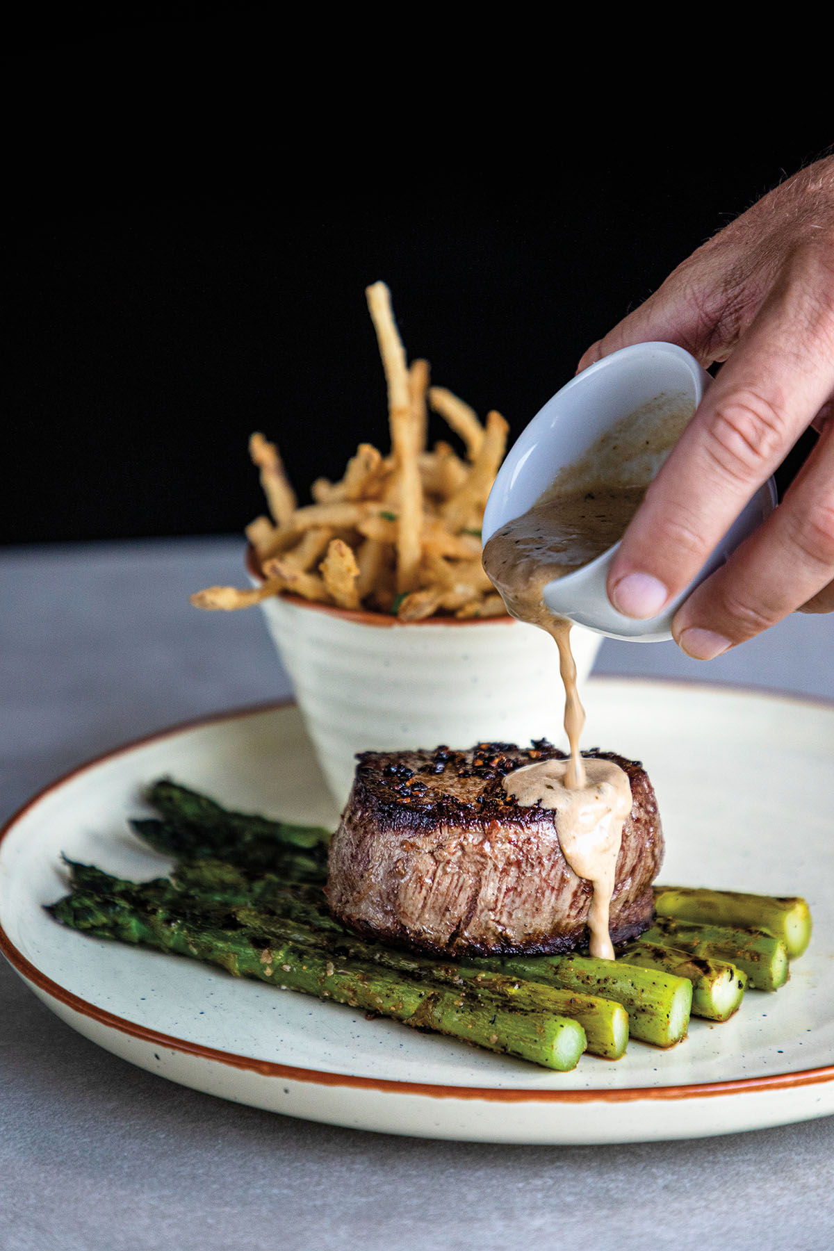 A hand holds a small dish of creamy sauce being poured over a grilled fillet mignon steak over asparagus with a side of French fries
