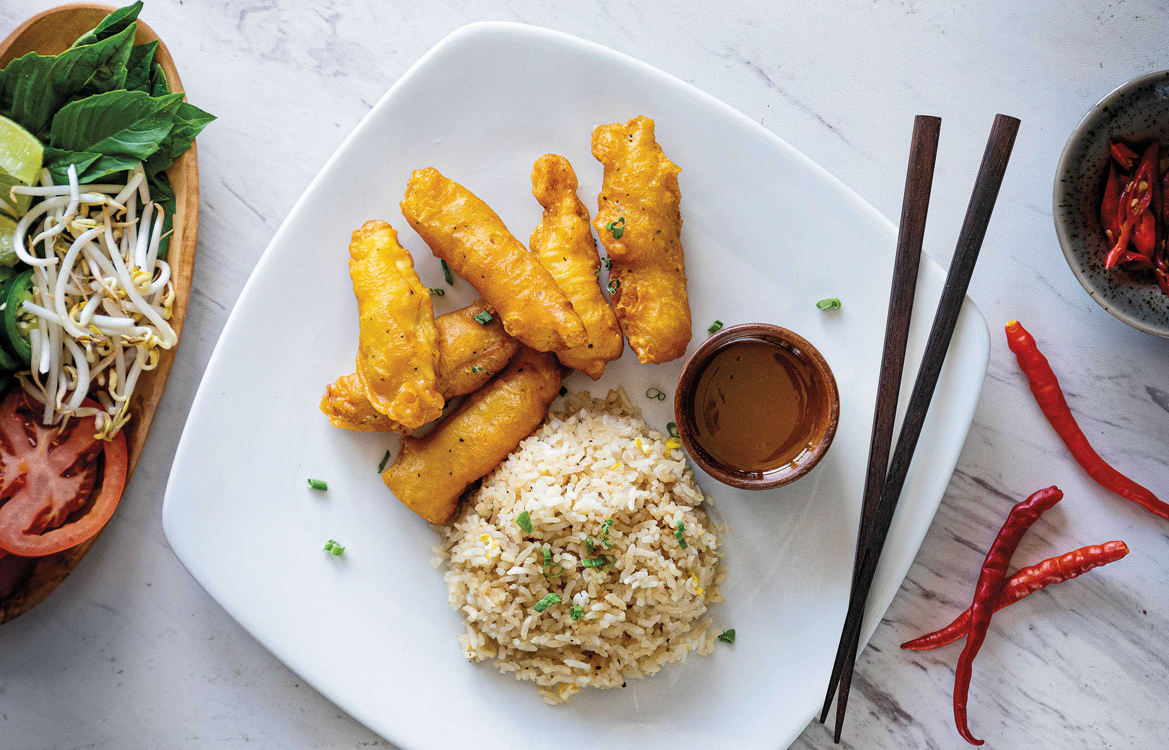 An overhead view of a white plate with a small circle of fried rice, a silver container of sauce, and a pile of golden-fried items next to chopsticks