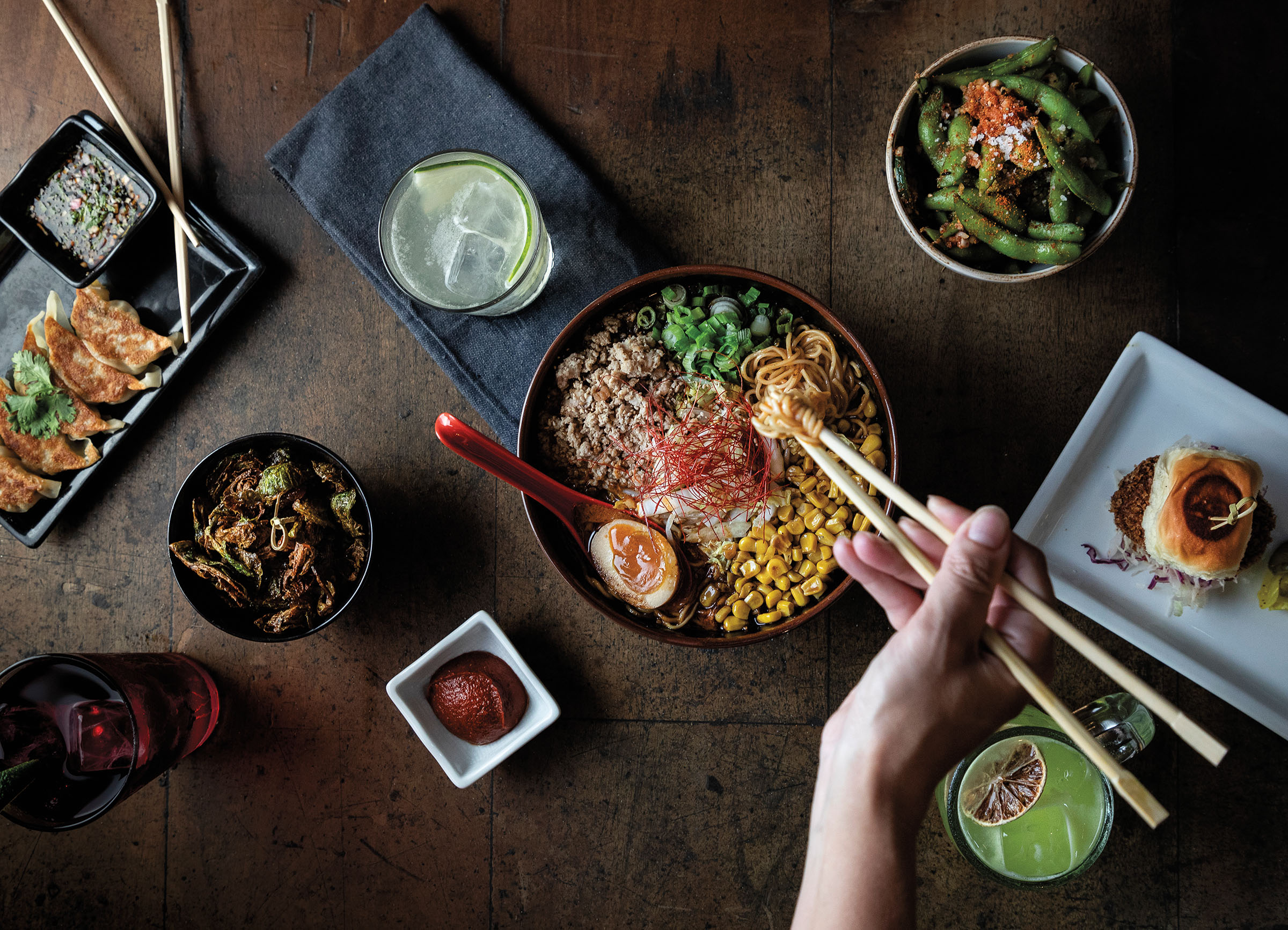 An overhead view of chopsticks dipping into a bowl of ramen, surrounded by various toppings and sides on a dark wood background