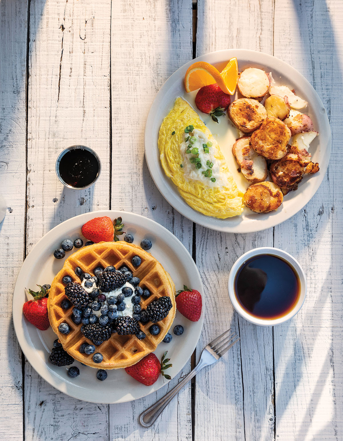 An overhead view of a light wood table with a waffle and berries on a plate, and another plate with an omlette and potatoes