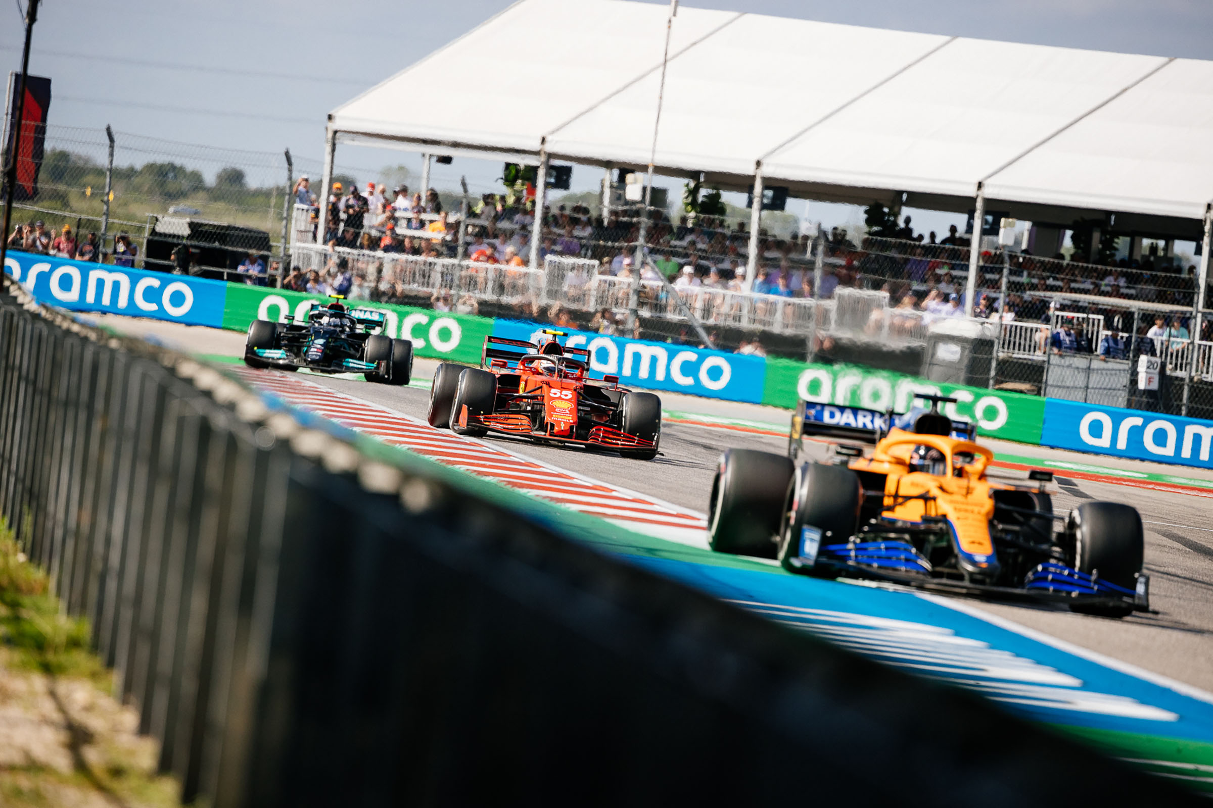 A yellow, red and green car are close as they enter the final turn on a racetrack with blue and green banners in the background reading 'aramco'