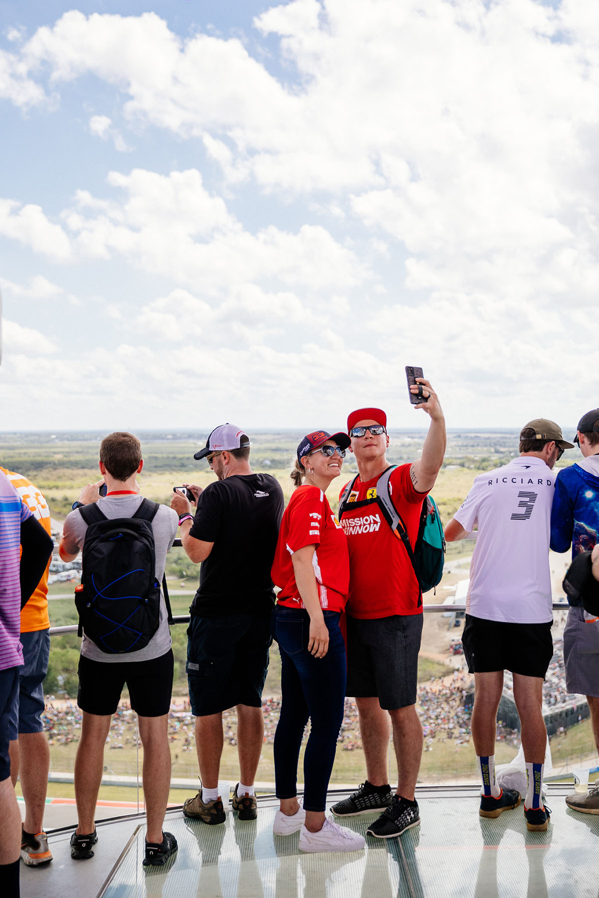 Two fans dressed in red and white pose for a selfie high in the air, with green grass, sky and clouds behind them