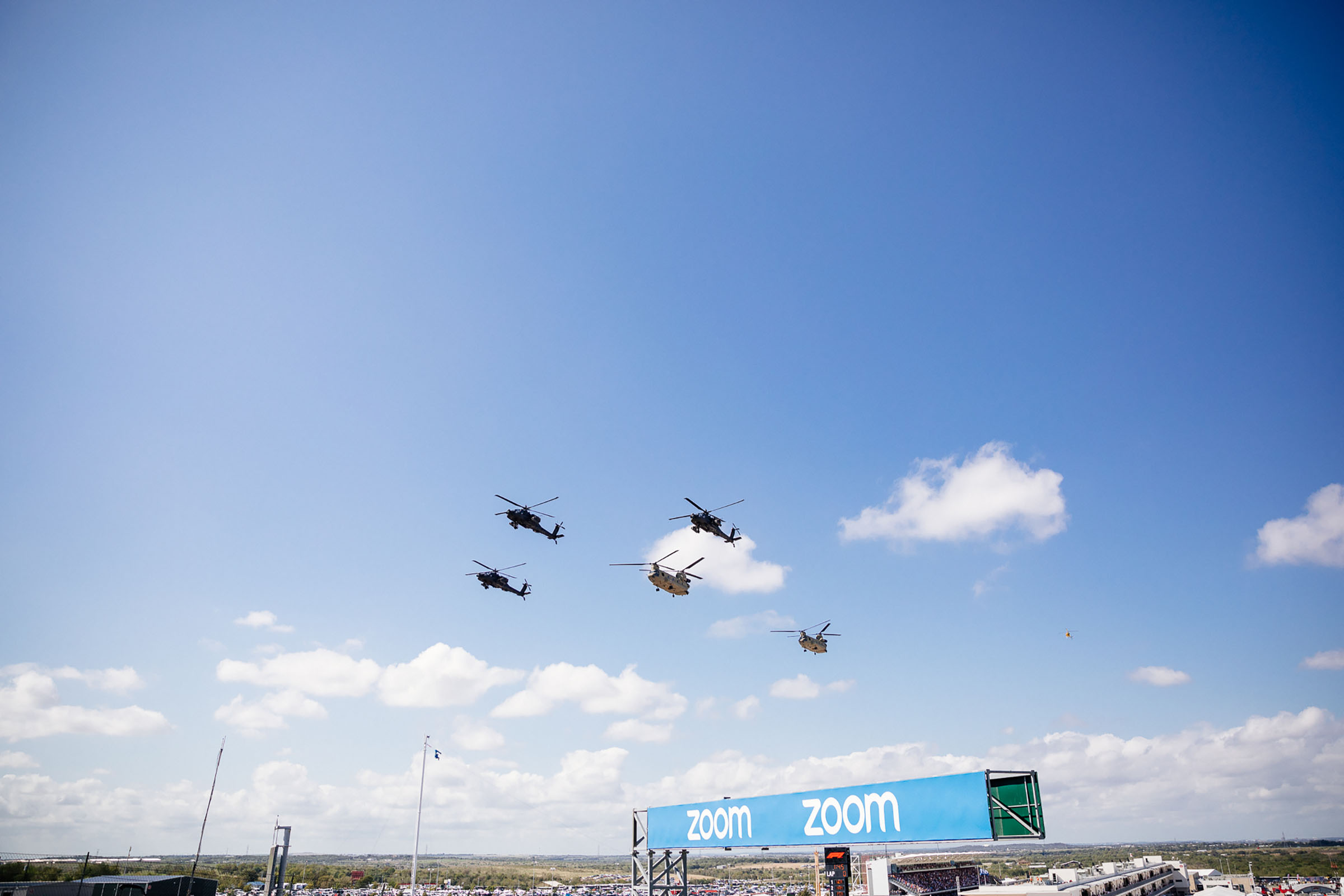 A group of five military helicopters fly over a banner reading 'Zoom' under blue sky