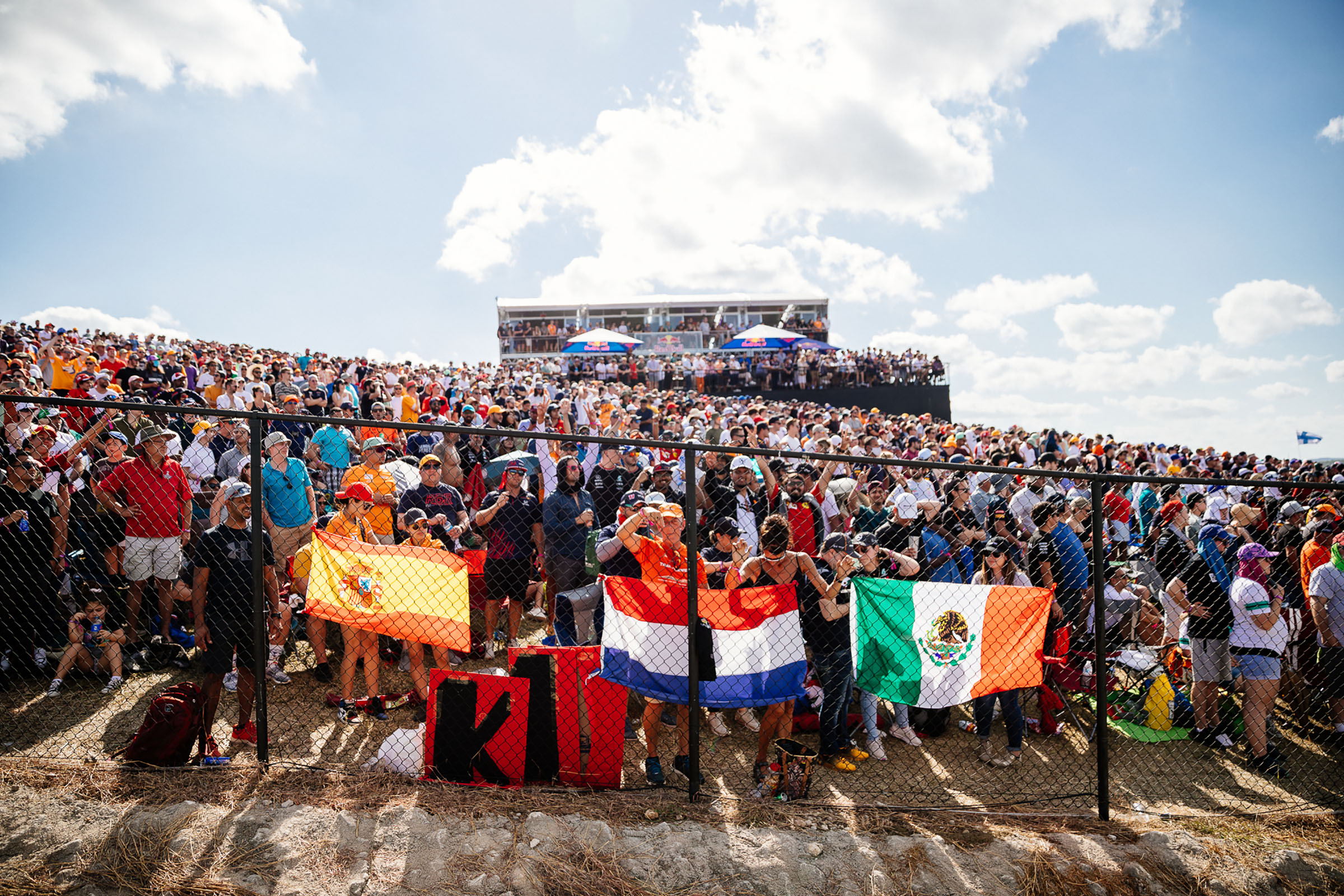Fans stand with flags of different countries behind a black fence in a large crowd of people