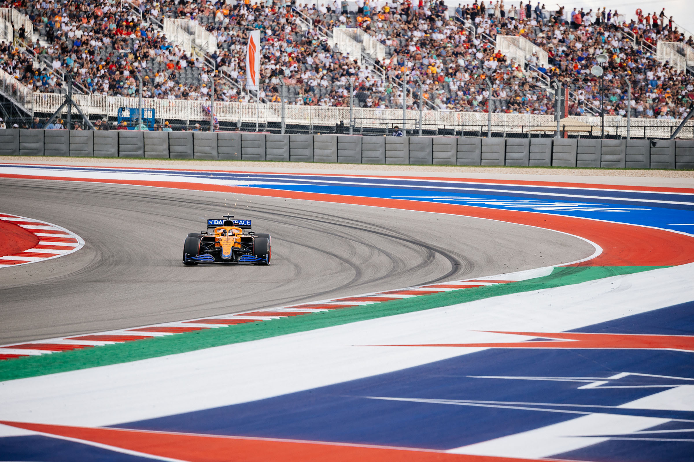 Sparks fly from behind an orange and blue car as it makes its way around a red, white and blue track