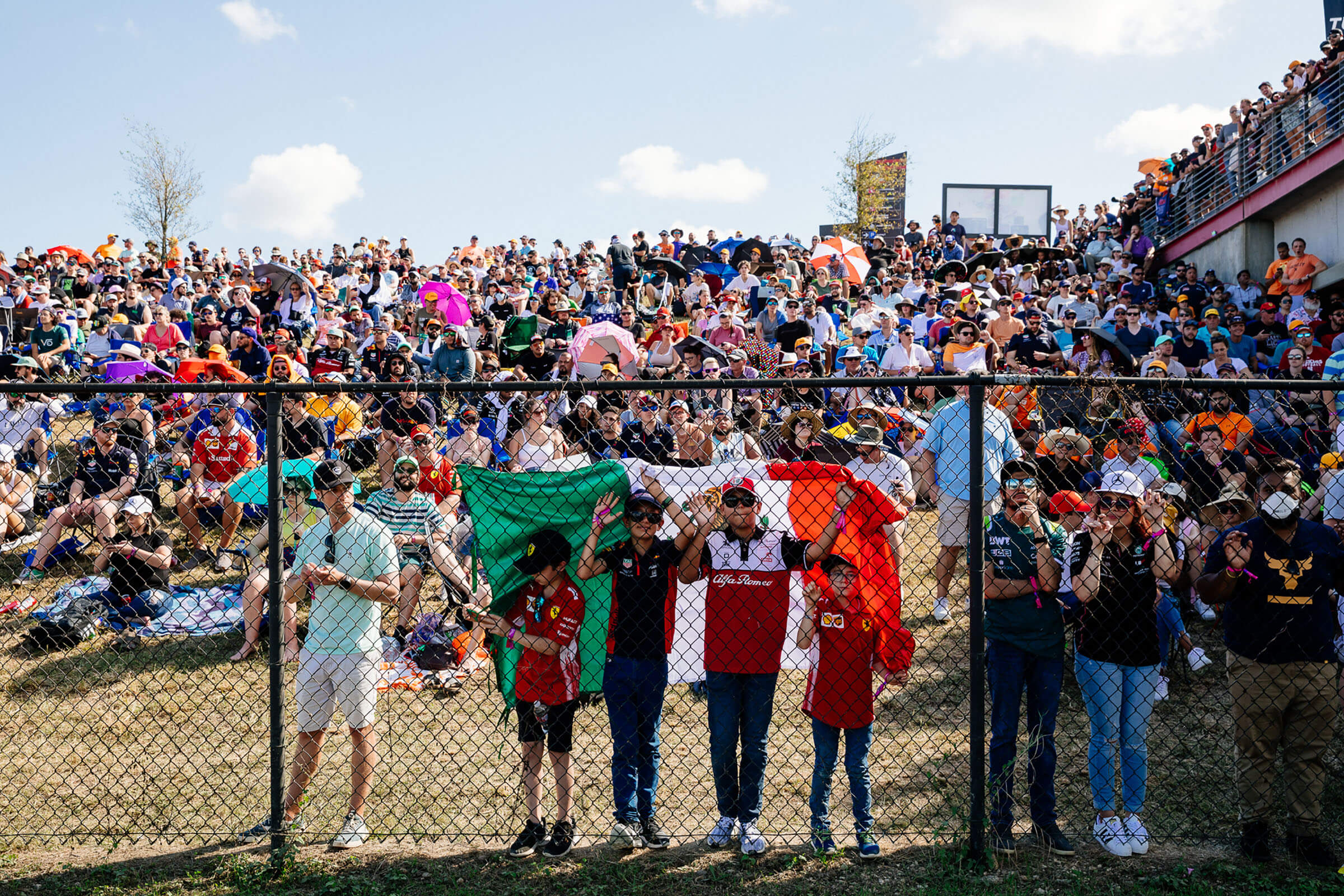 A group of people stand under the flag of Mexico amid large crowds of people sitting on grass