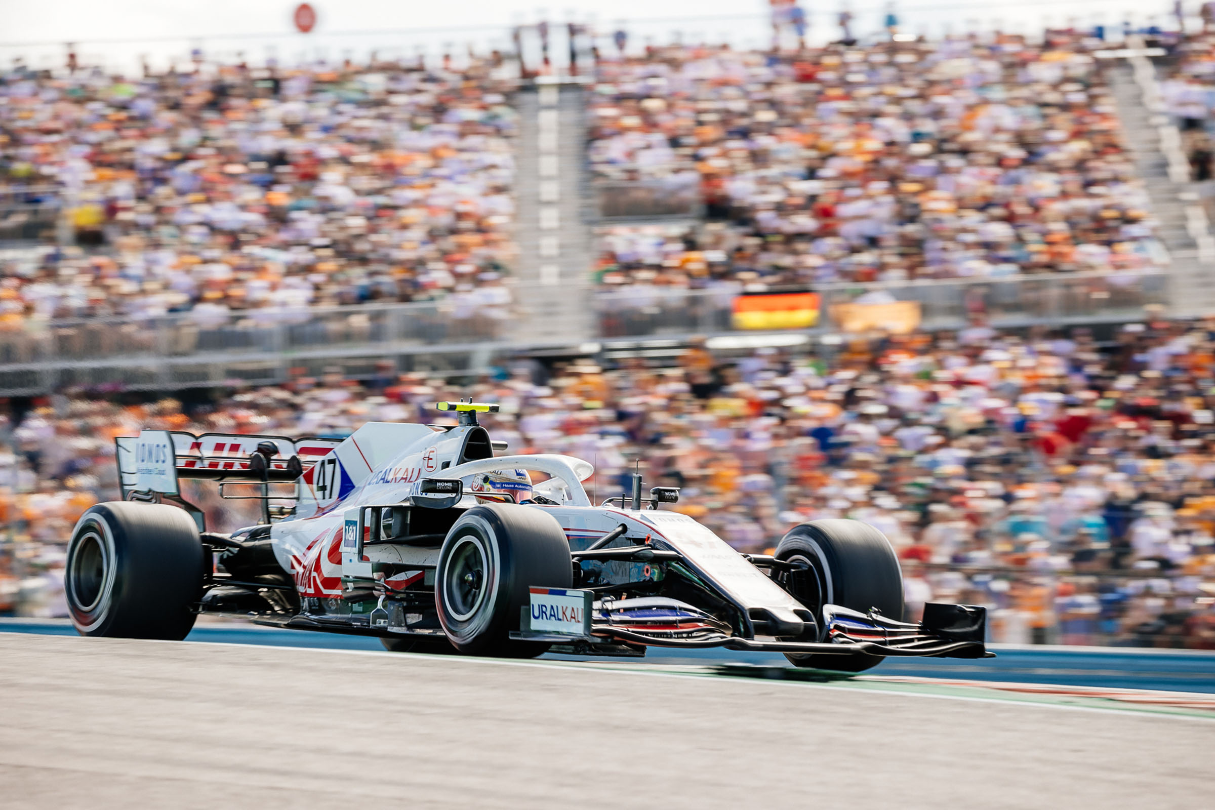 A gray car with large black tires speeds in front of a large crowd of people