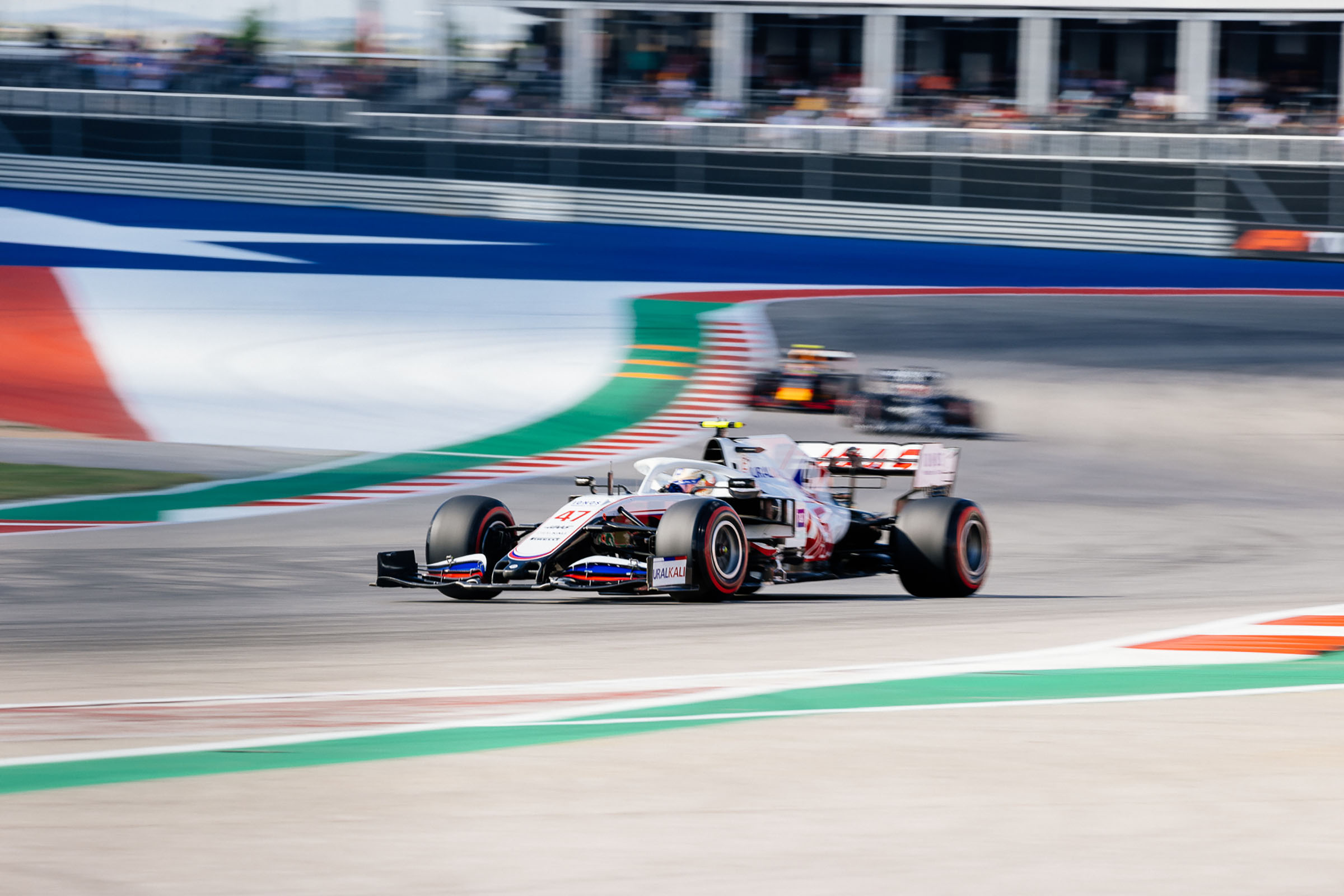 A red white and blue car rounds a corner with a Texas flag painted on the asphalt behind