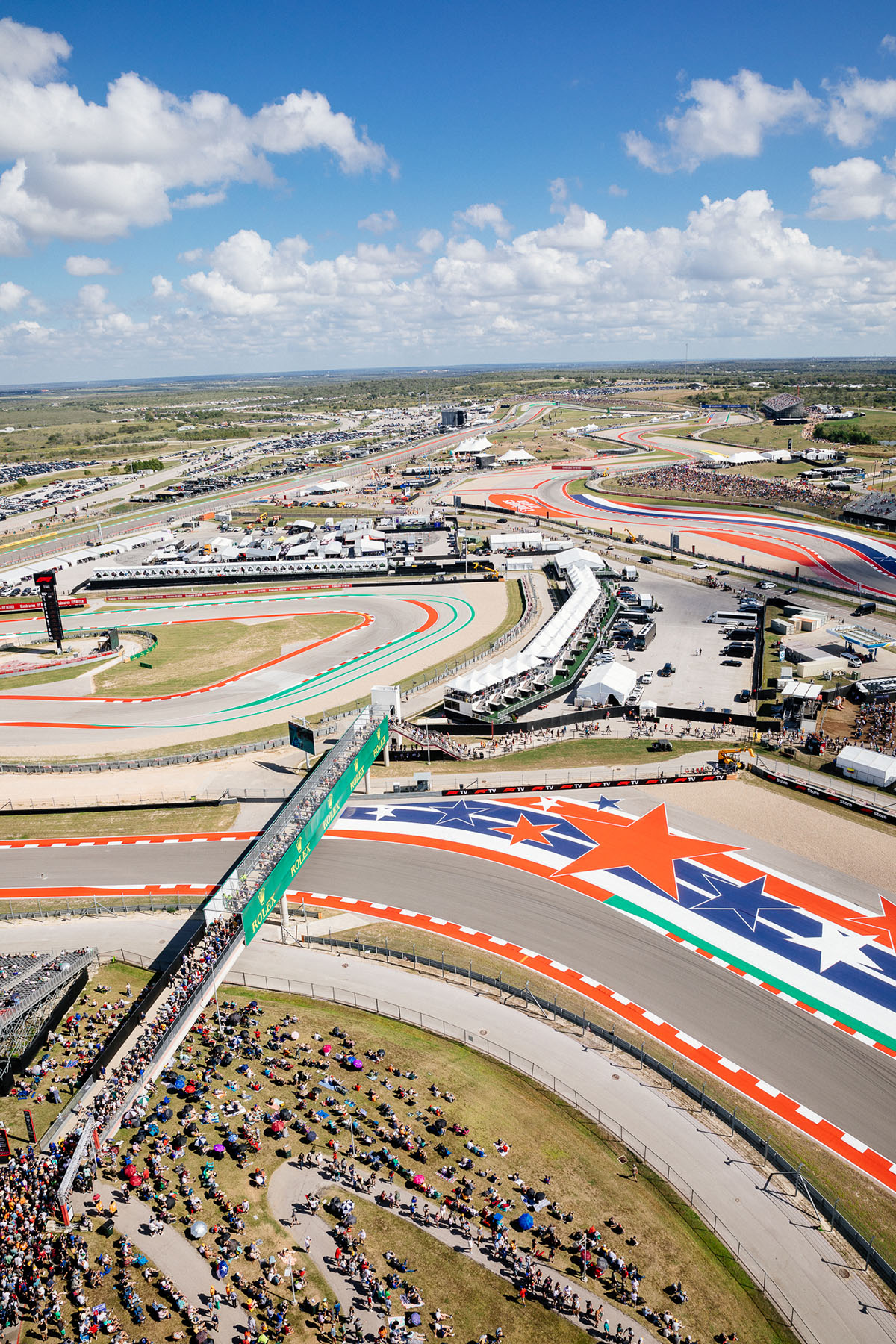 An overhead view of blue sky and green ground with many brightly-painted asphalt tracks and crowds of people