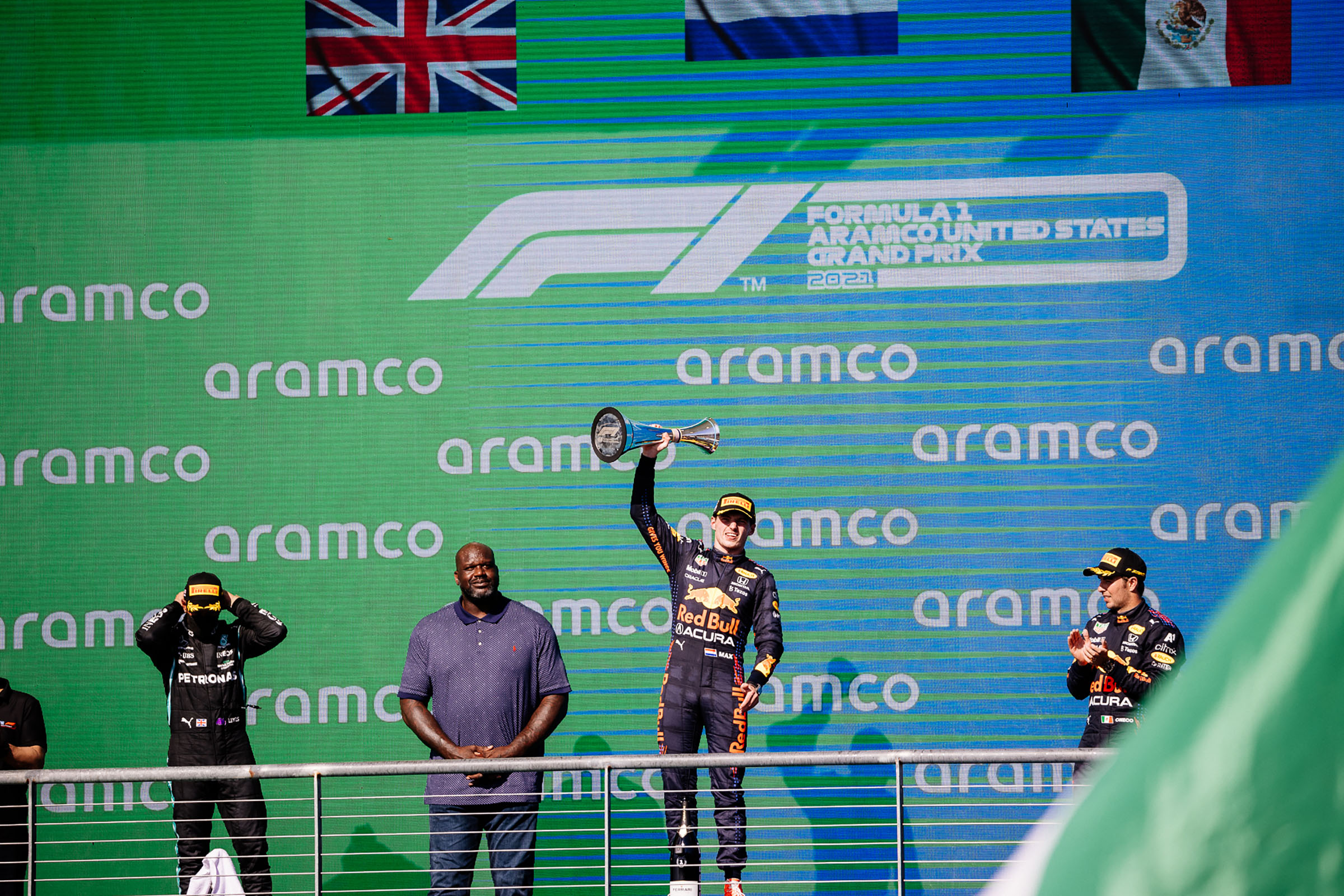 A man in a Red Bull uniform holds a gold trophy in his right hand next to a man in a blue polo shirt in front of a Formula 1 and aramco background