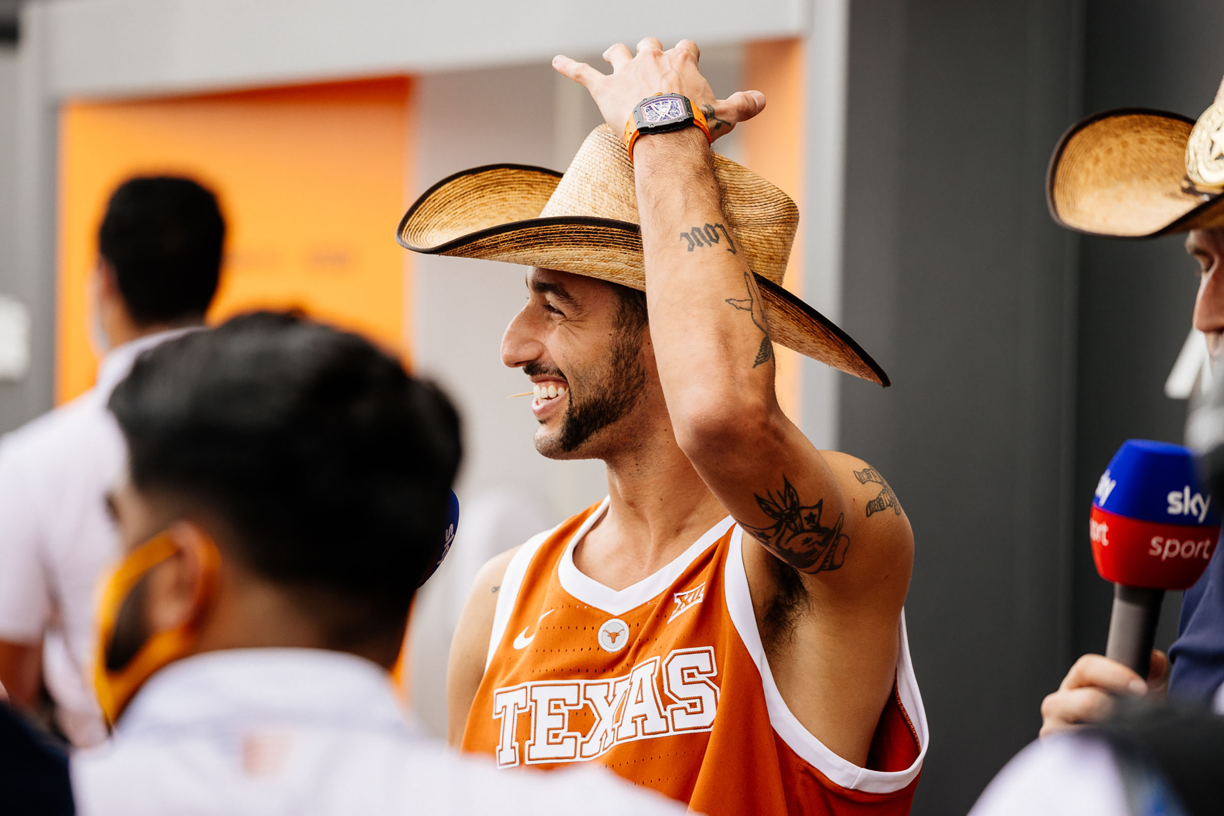 A man in a burt orange Longhorns jersey smiles while putting his hand near a straw cowboy hat on his head