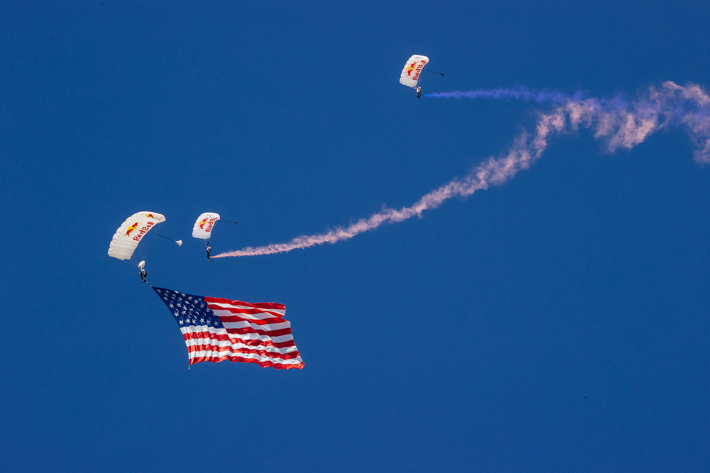 The American flag flies behind a group of three skydivers in 'Redbull' parachutes under red, white and blue smoke